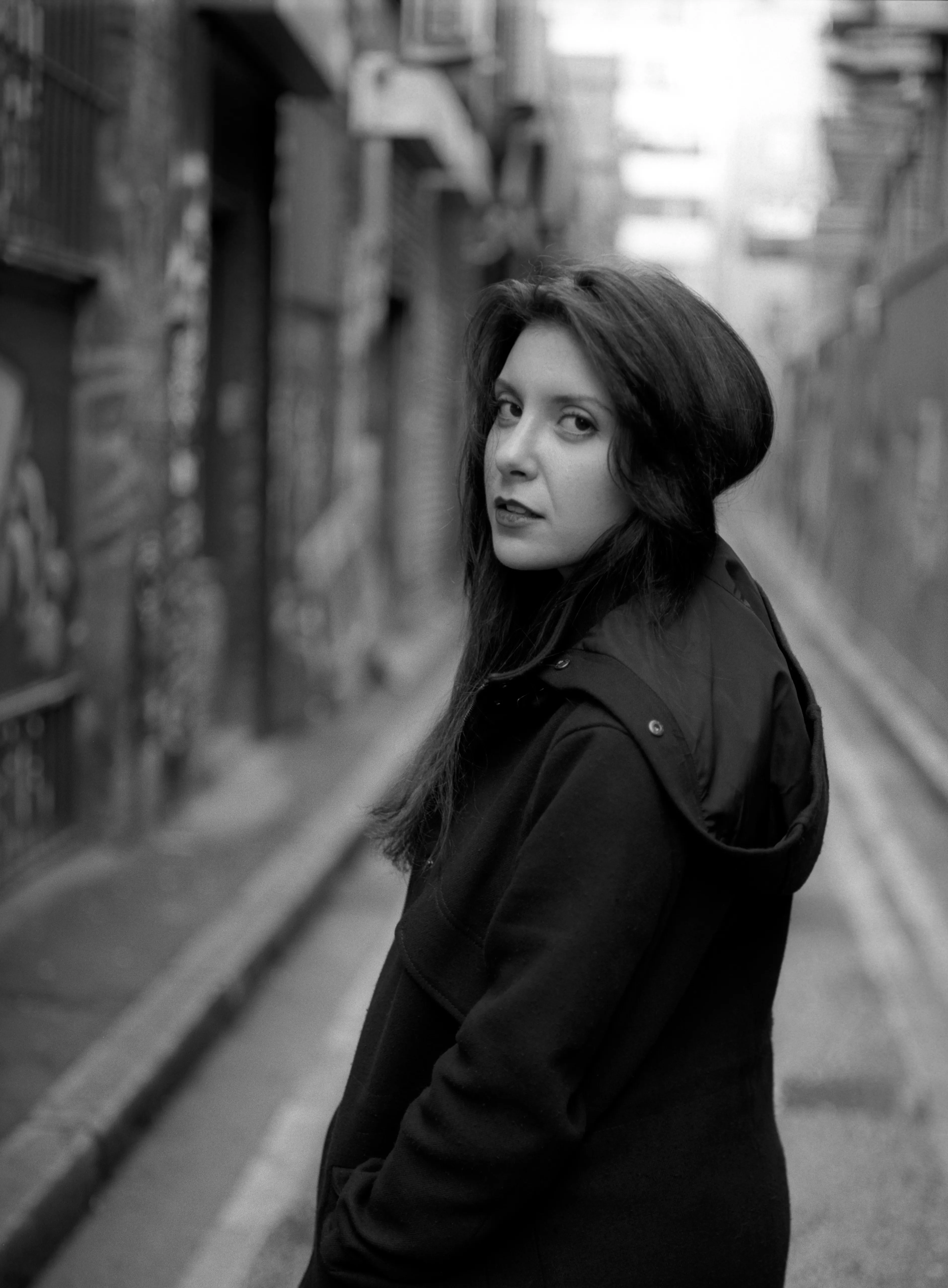 A woman in a dark hooded coat looks back over her shoulder in a narrow, graffiti-lined alley that recedes behind her.