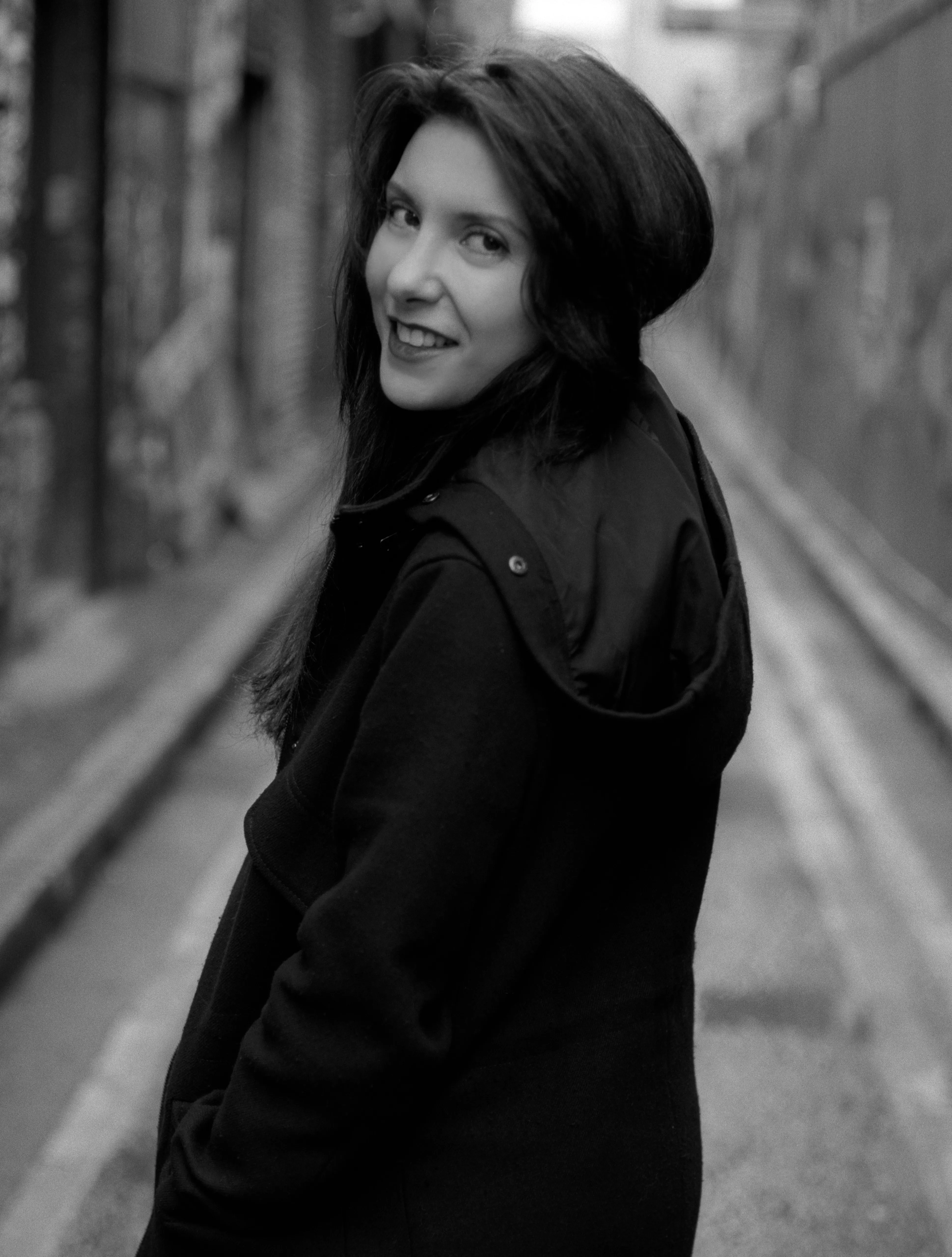 A woman turns back over her shoulder with a broad smile in a narrow, graffiti-covered alley.