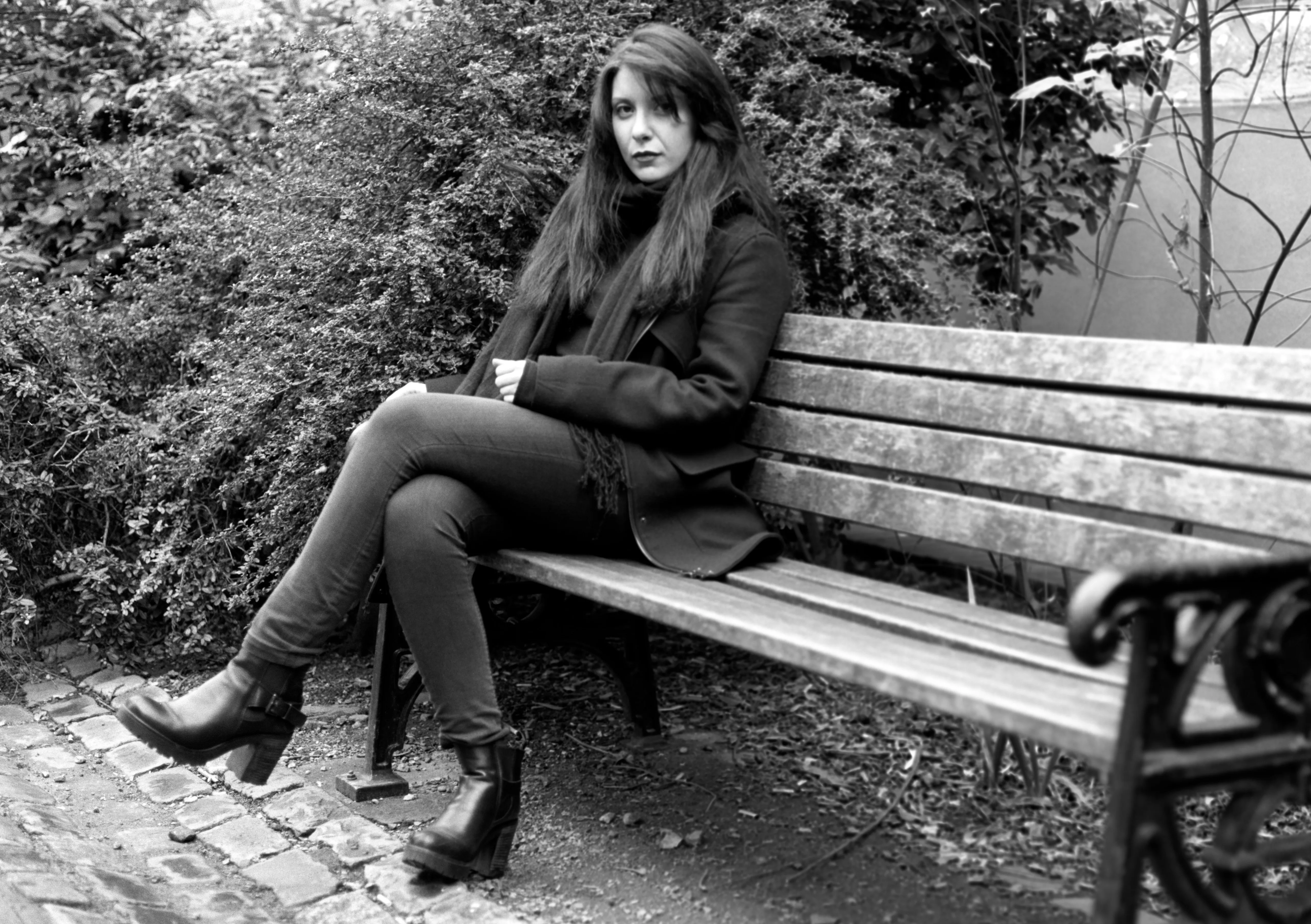 A woman sits at one end of an empty park bench, hands folded, surrounded by foliage.