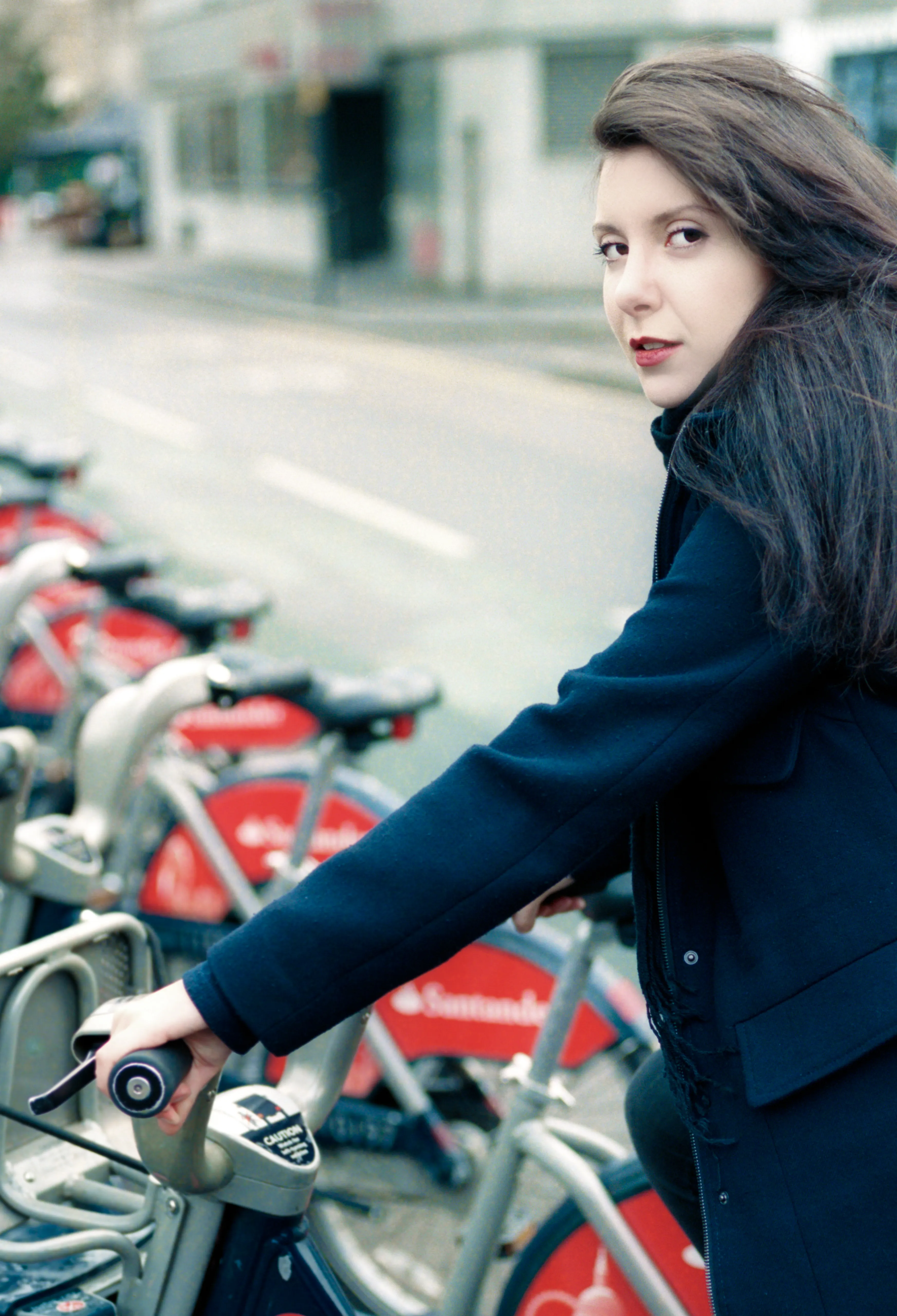 A woman in a dark coat stands beside a row of red hire bikes on a street, glancing back over her shoulder.