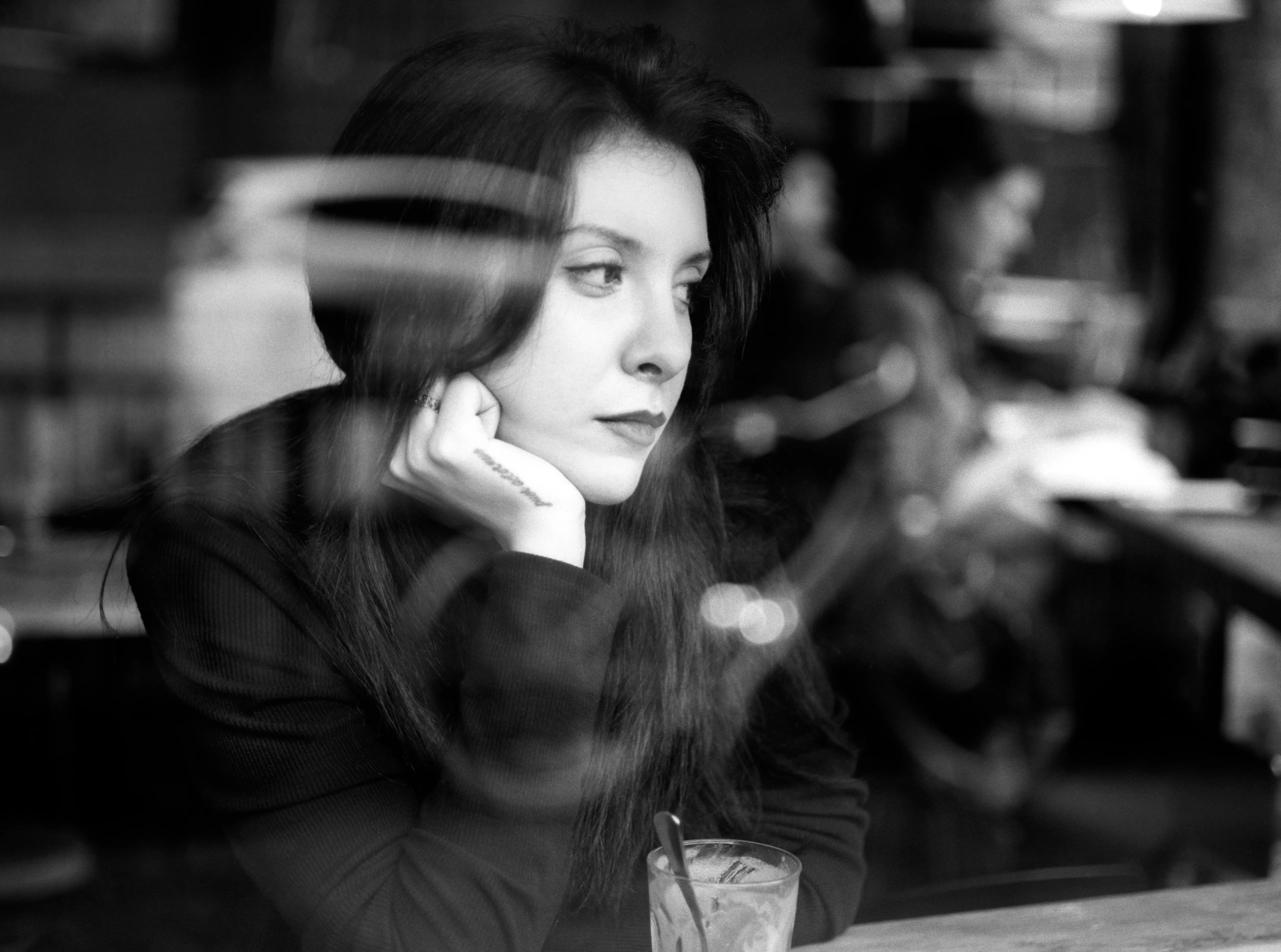 A woman sits at a café table with her chin on her hand, gazing past the camera; a blurred streak of light crosses the frame behind her.