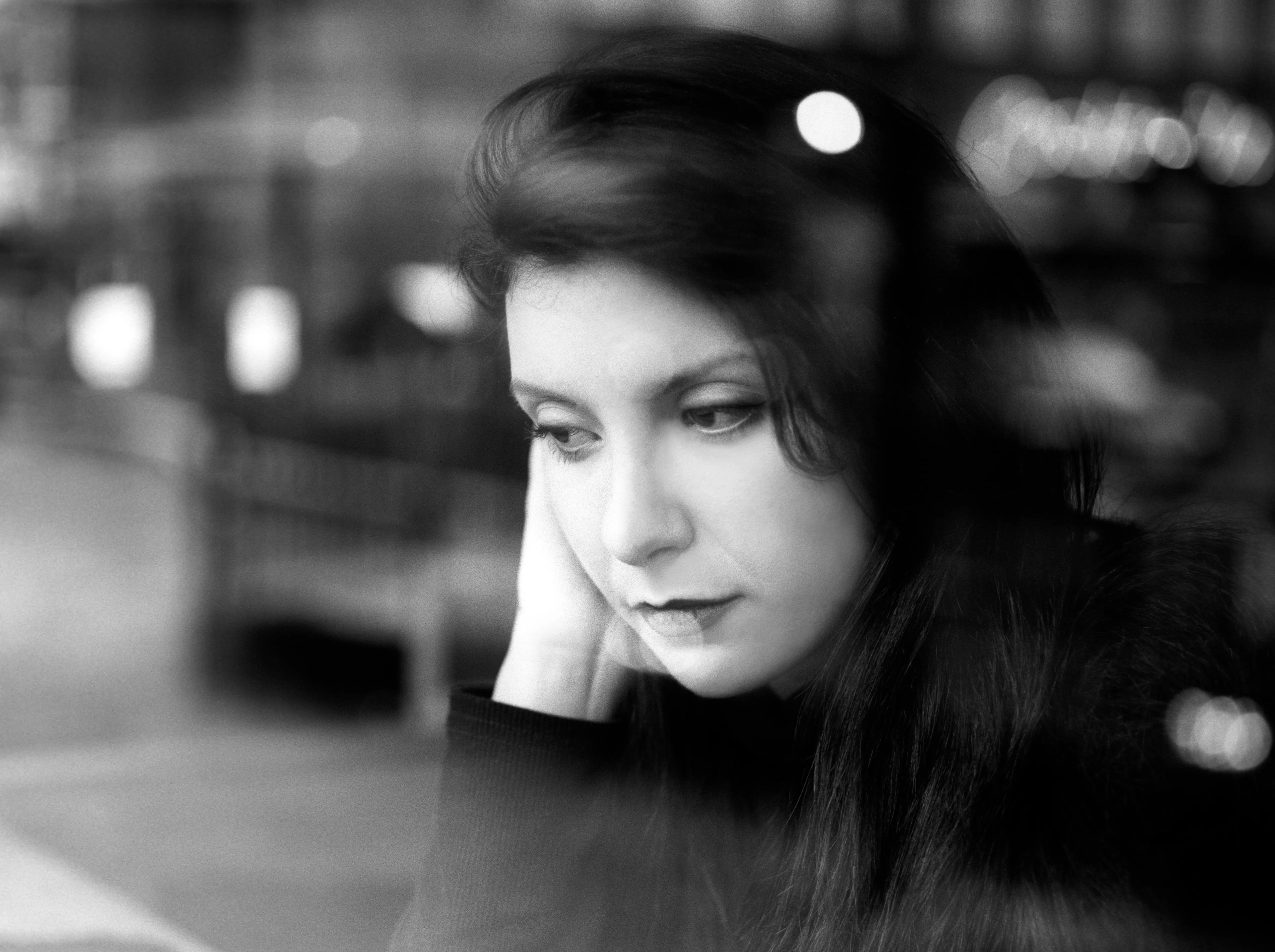 A woman rests her cheek on her hand with her eyes lowered, against softly blurred evening city lights.