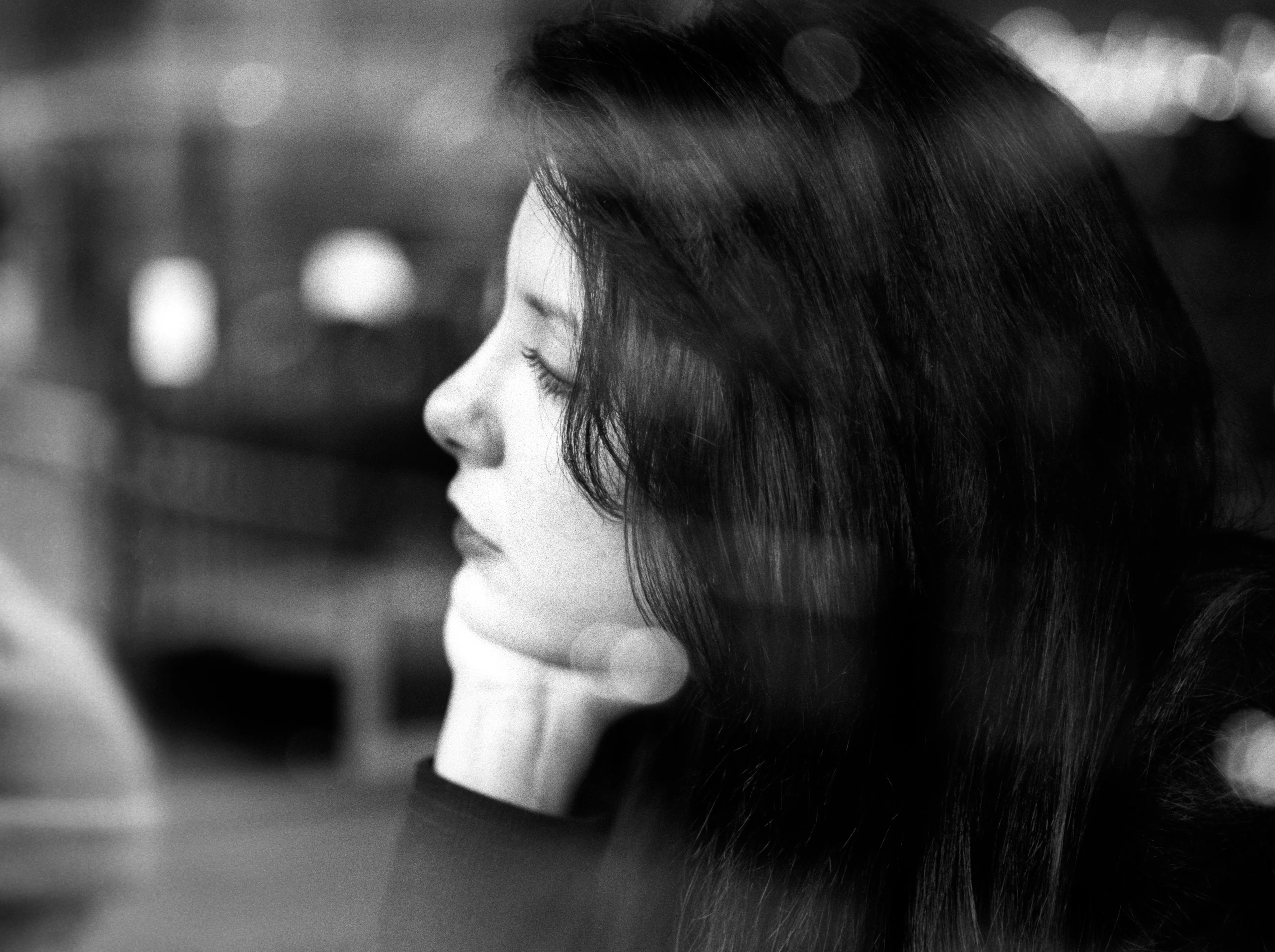 Profile of a woman with her eyes closed and a hand beneath her chin, blurred city lights behind.