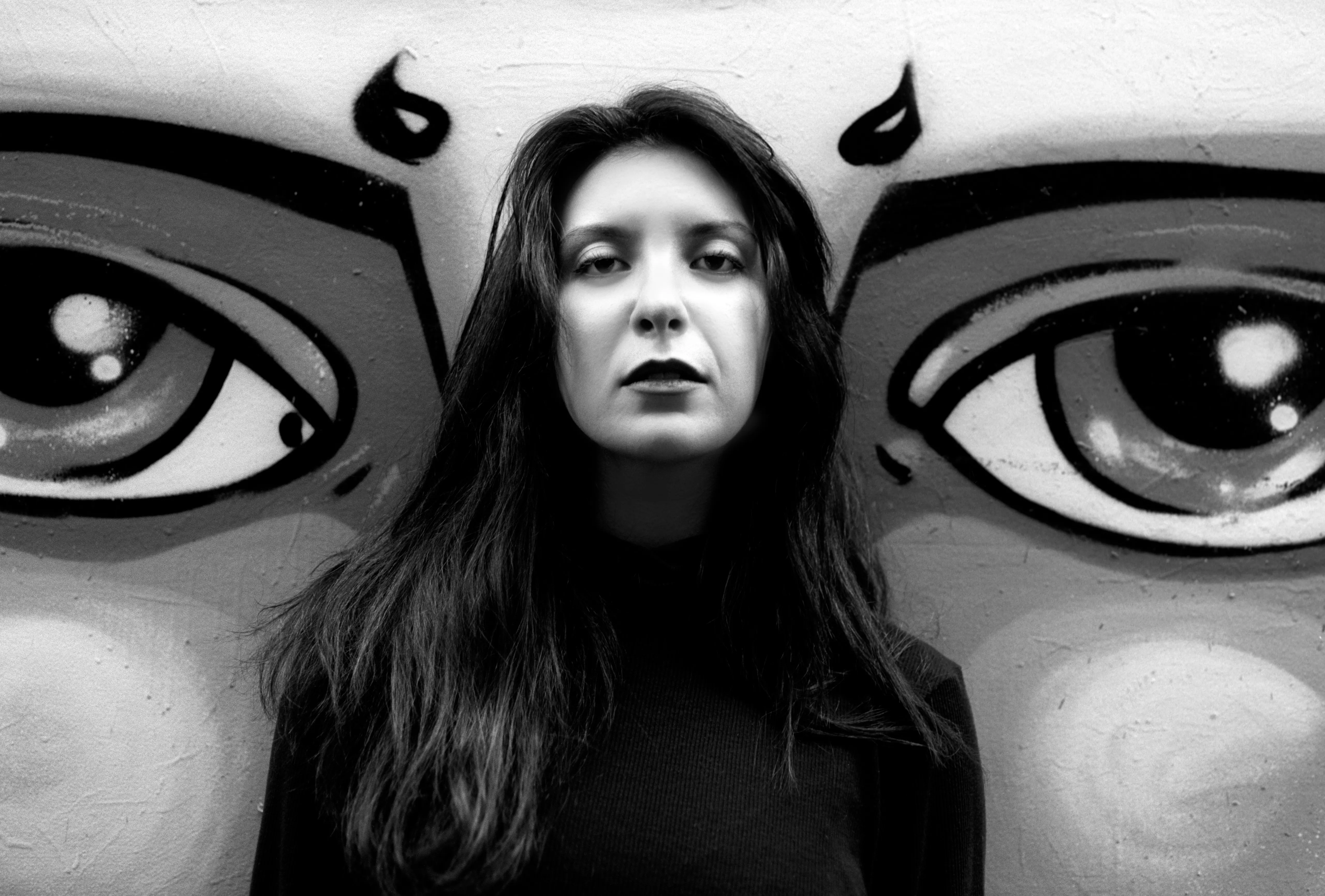 A woman stands against a wall painted with two large eyes, her face centred between them as she looks at the camera.