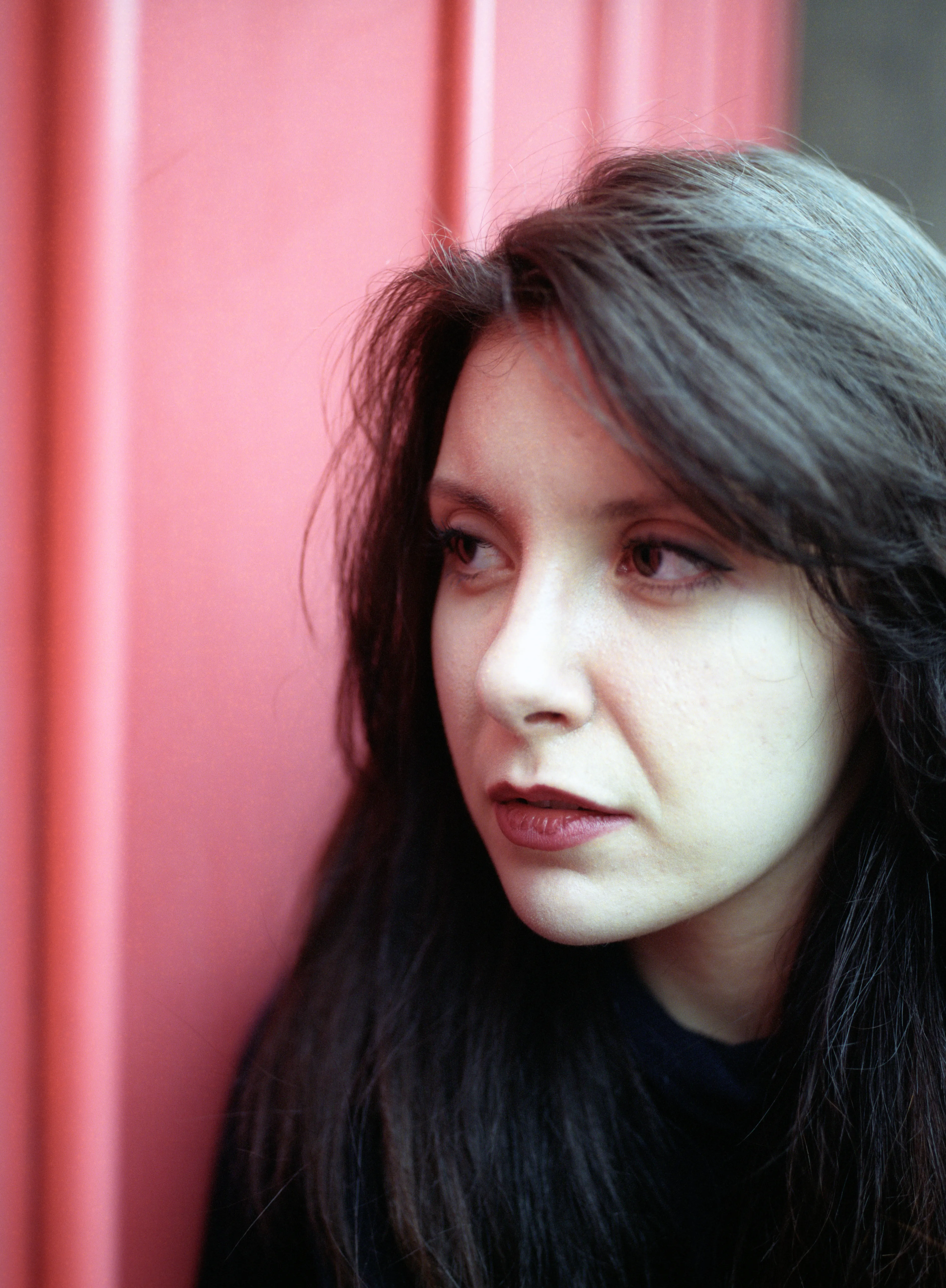 A woman with long dark hair leans against a coral-pink panelled door, turned slightly away and looking to her left.