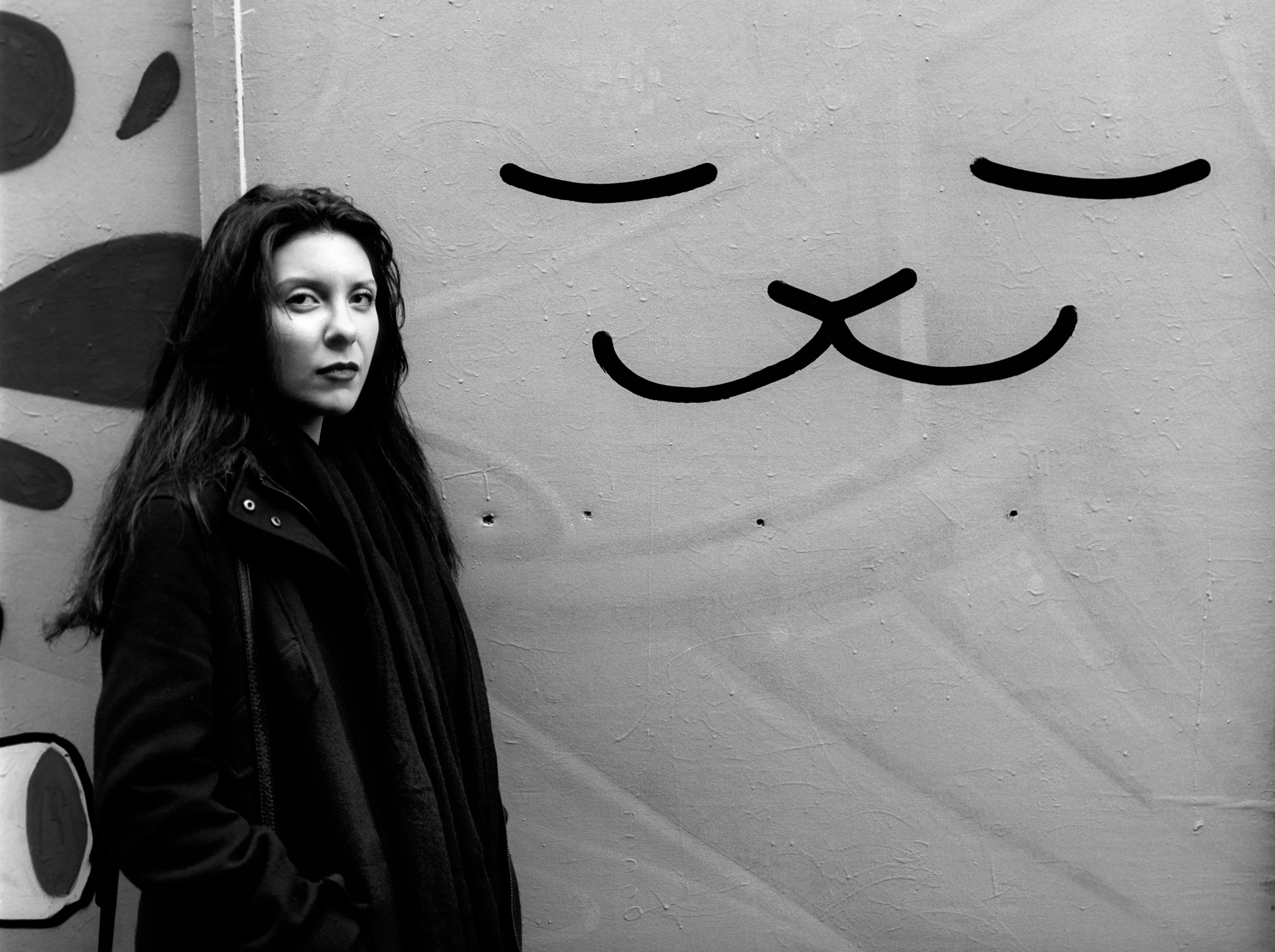 A woman in a dark coat stands to one side of a pale wall marked with two large painted, smile-like curves.