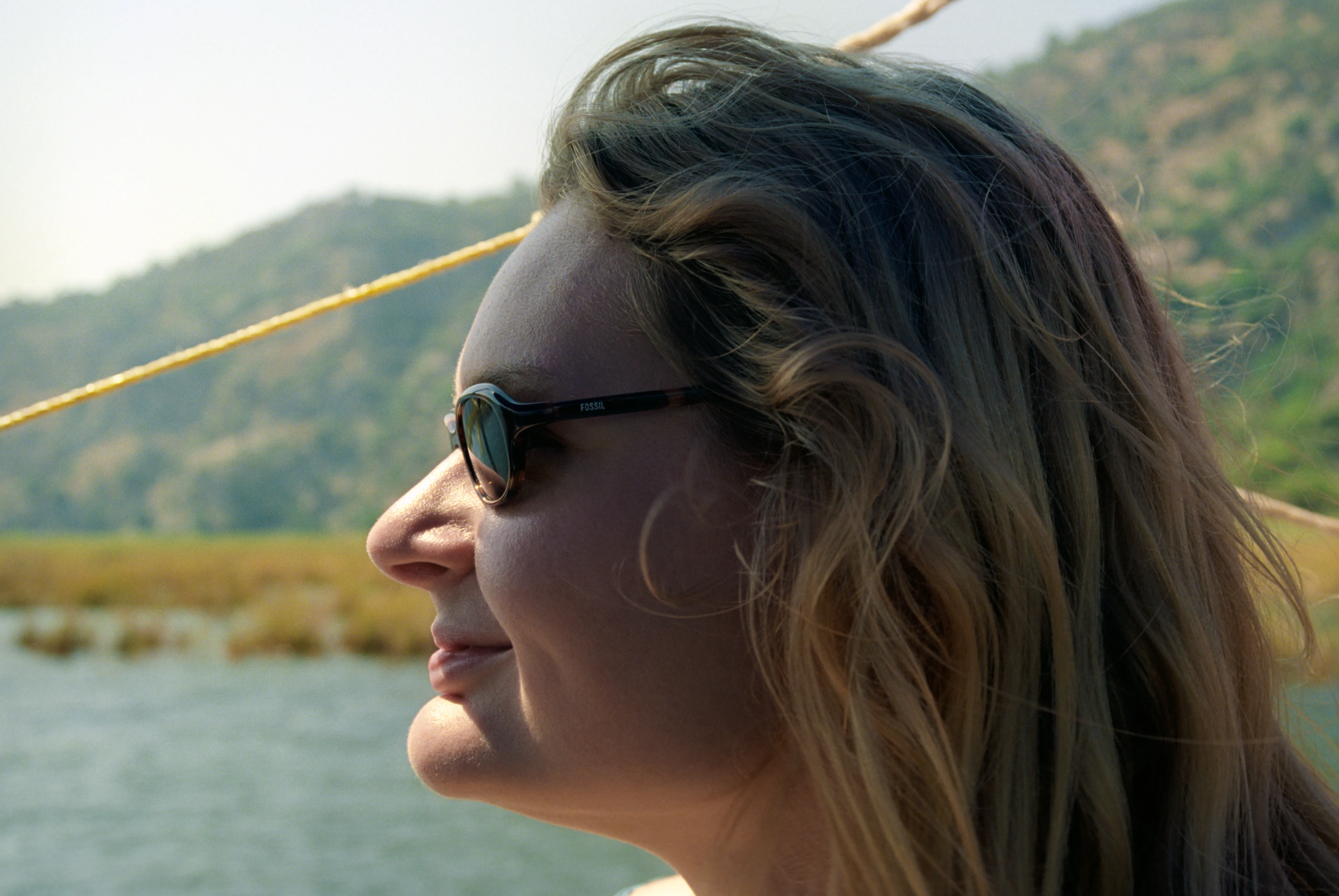 Liddy in profile, wearing sunglasses, lit by late sun on a gulet, with a rigging line crossing the frame and Turkish hillsides behind.