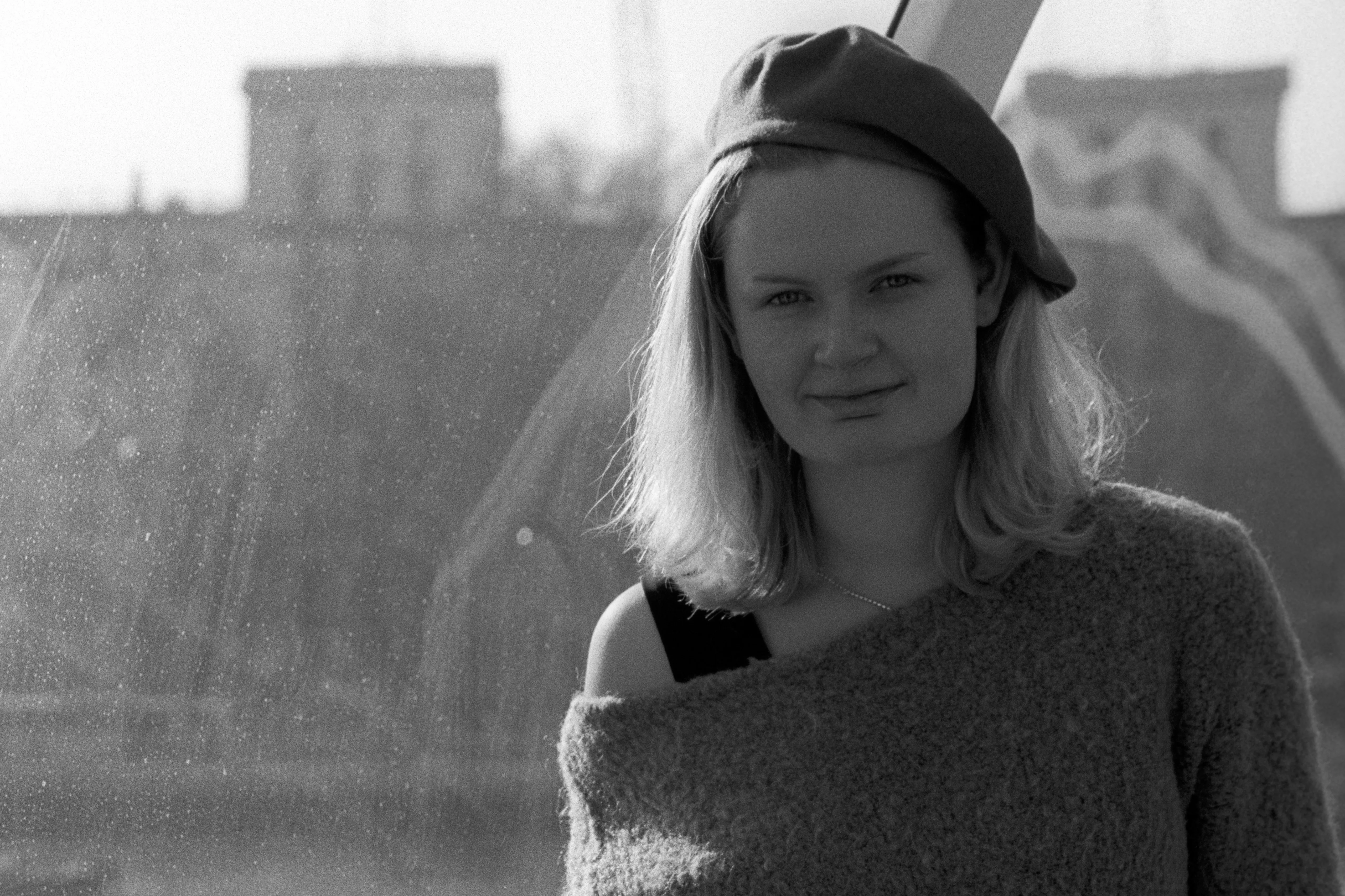 Liddy in a beret and off-shoulder knitted sweater, half-smiling at the camera through a dust-marked window, industrial rooftops blurred behind.