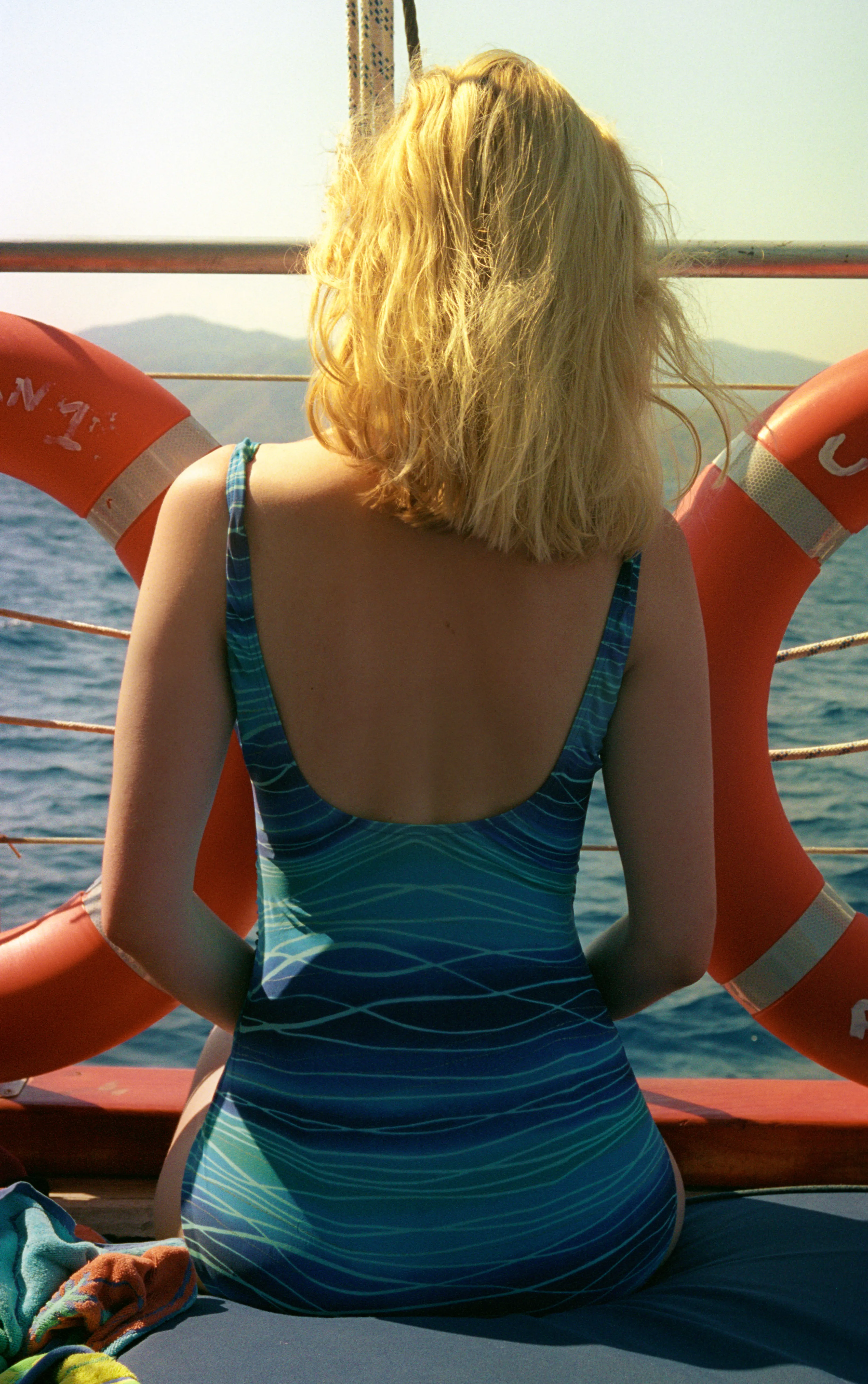 Liddy seen from behind sitting on a boat deck in a wave-pattern blue swimsuit, framed symmetrically between two orange life rings, looking out across the sea.
