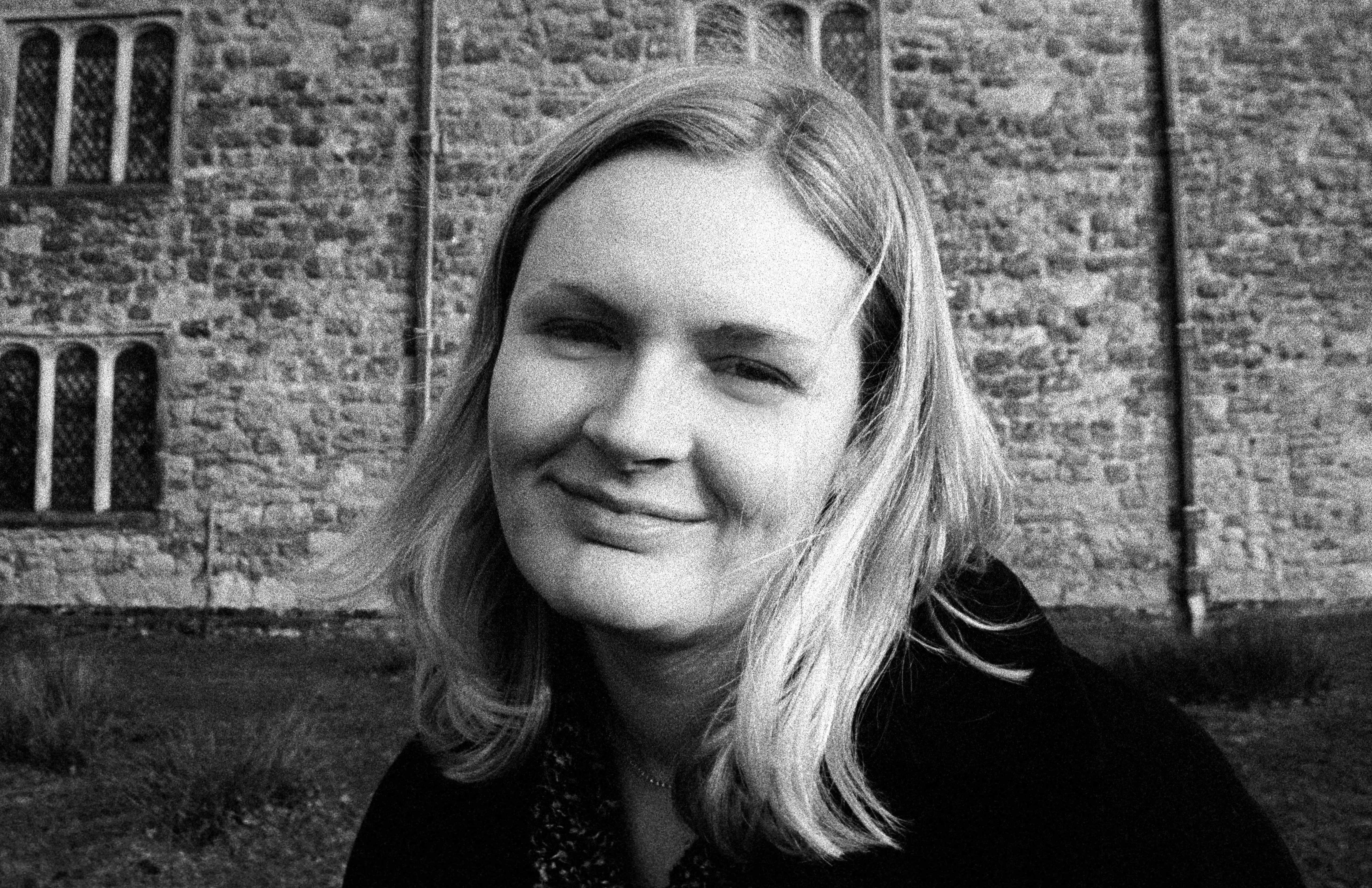 Liddy half-winking and smiling in front of a stone wall with leaded gothic windows. Heavy film grain.
