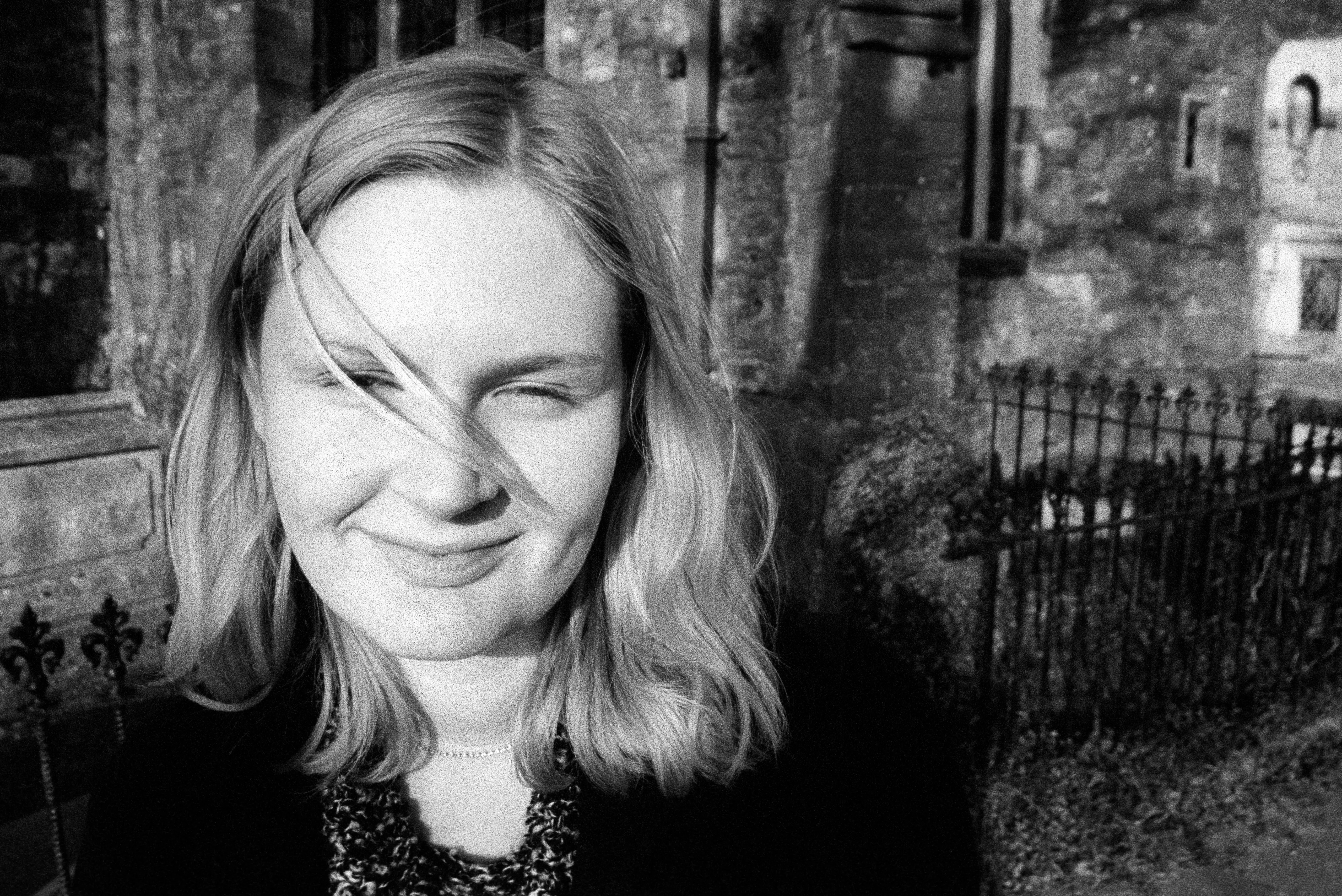 Close portrait of Liddy with hair blown across her face, one eye half-closed, smiling. Gothic stone wall and iron railing behind.