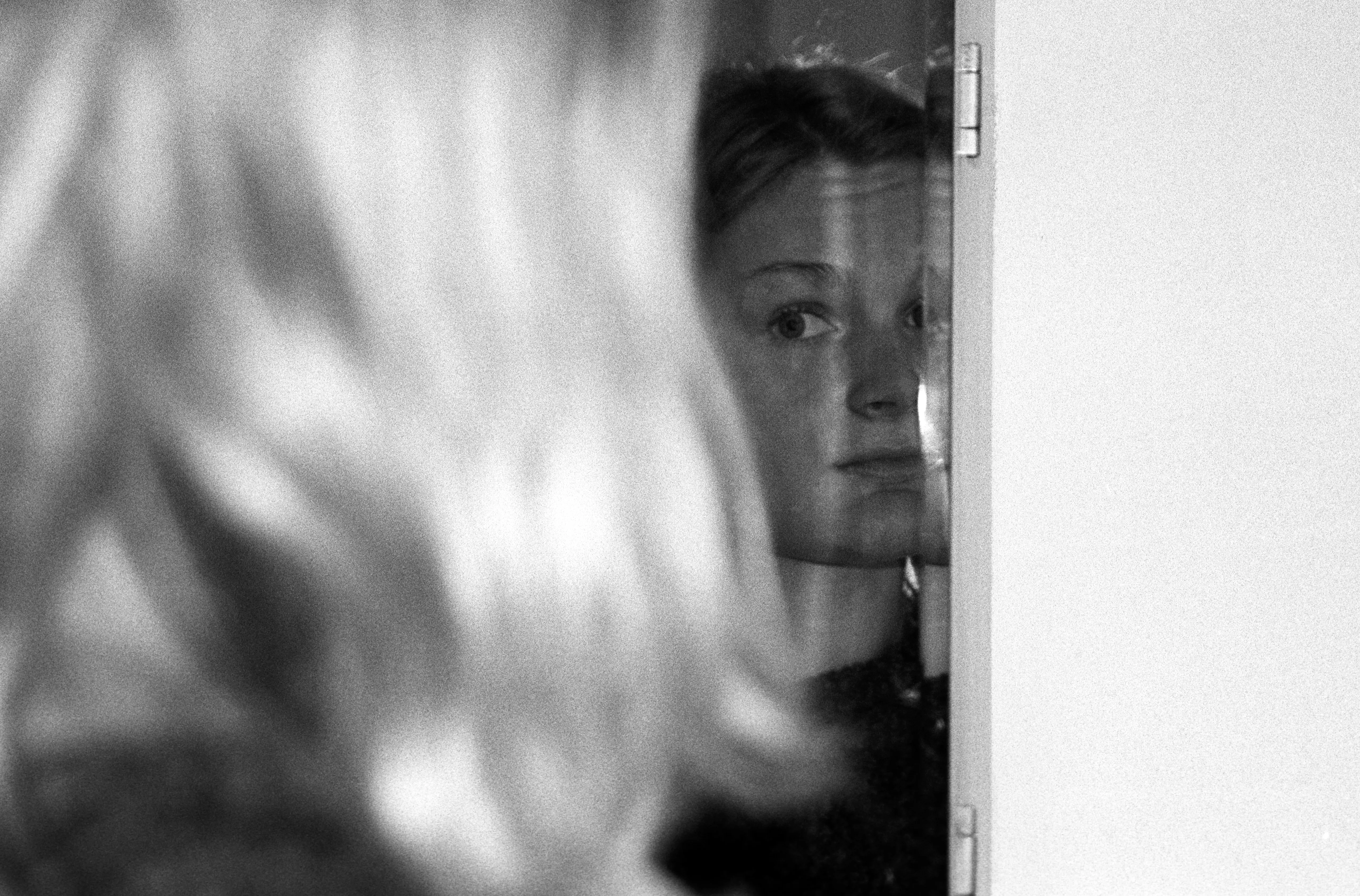Liddy's face seen through a pane of glass, foreground hair blurred across the left of the frame, her eyes meeting the camera obliquely.
