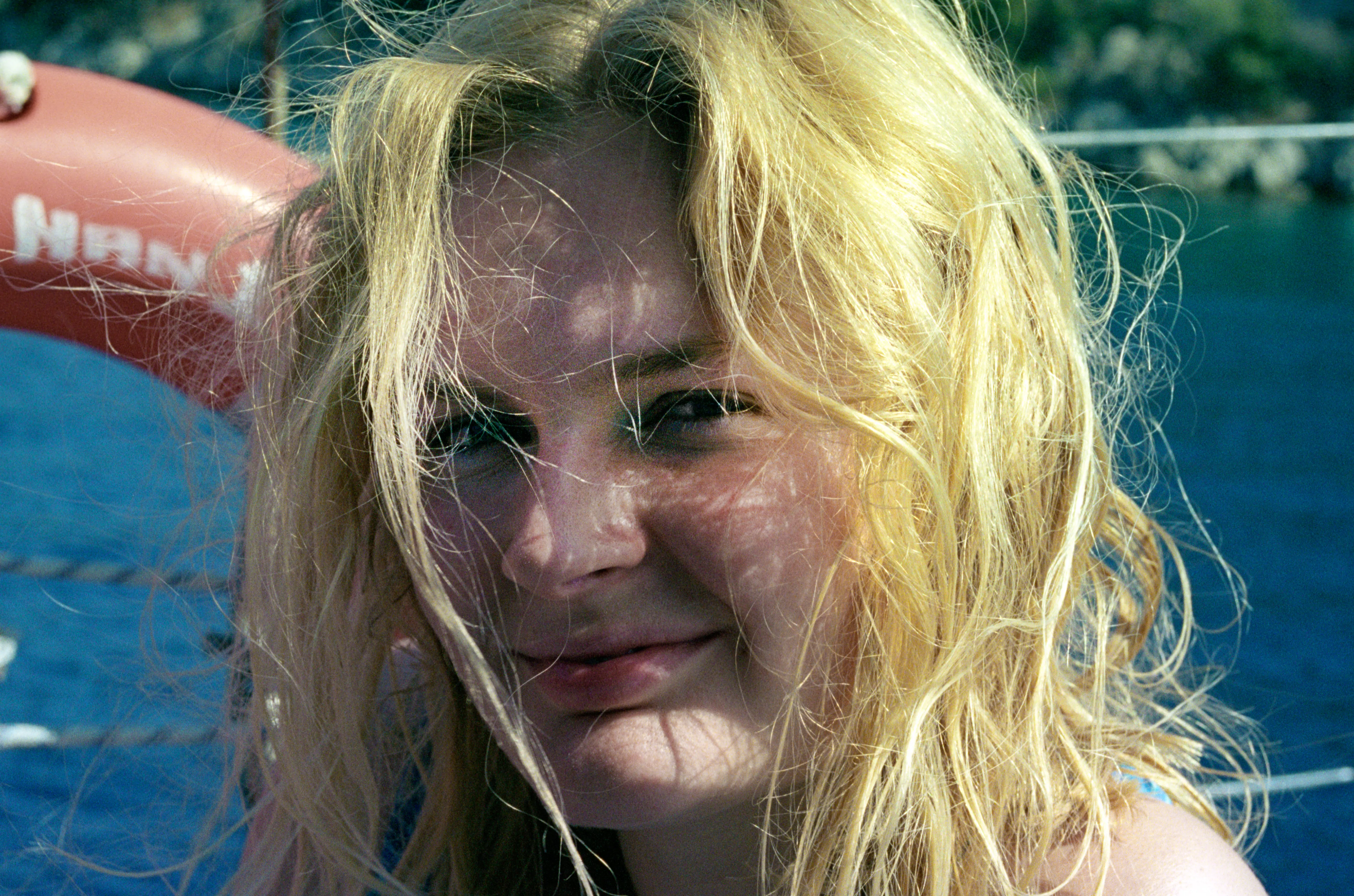 Liddy on a boat, hair wind-tossed and golden in strong sun, dappled shadow falling across her face, a red life ring visible behind her.