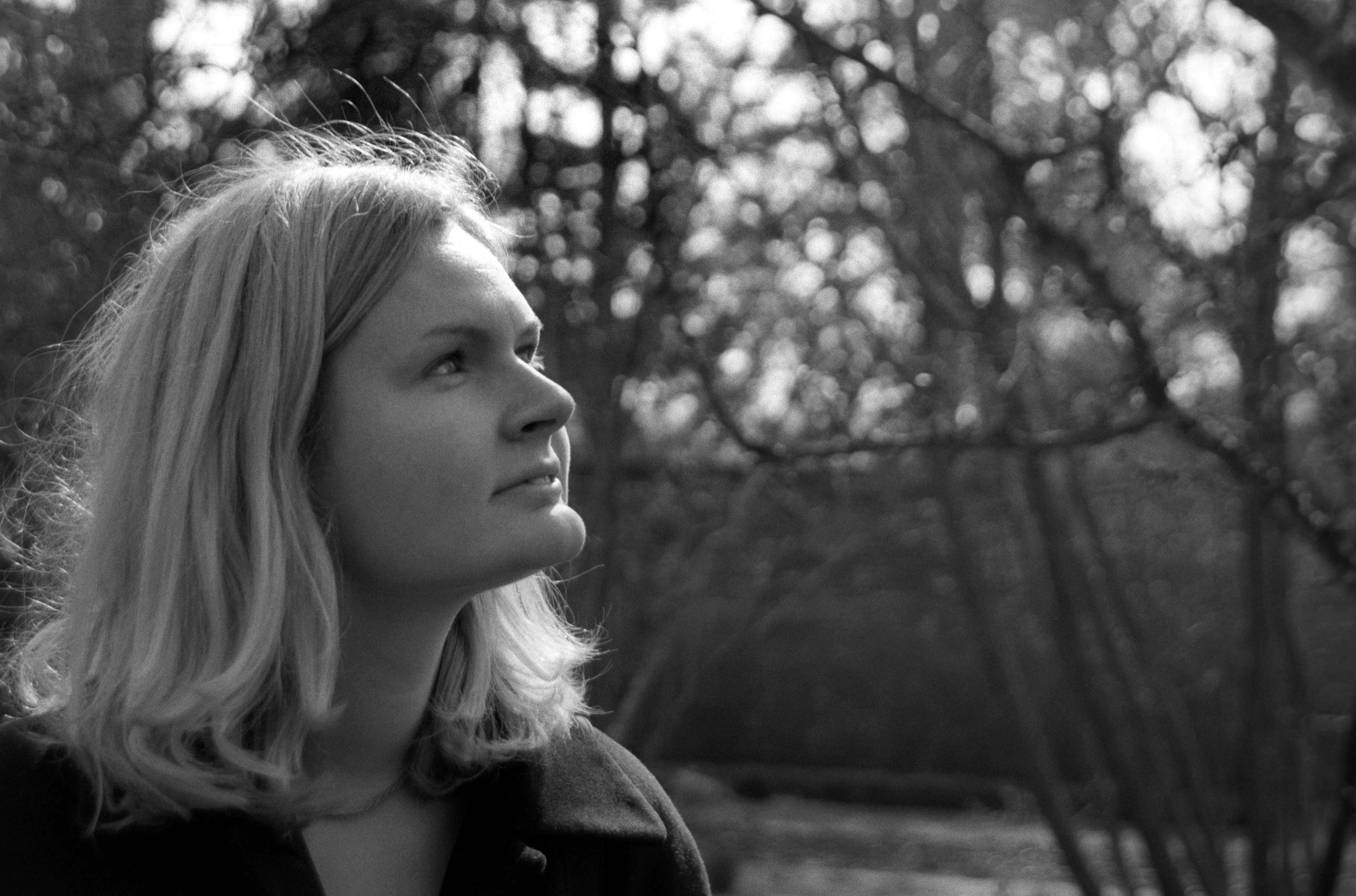 Liddy in three-quarter view looking up into the light, hair lit from above, dappled tree bokeh behind her.