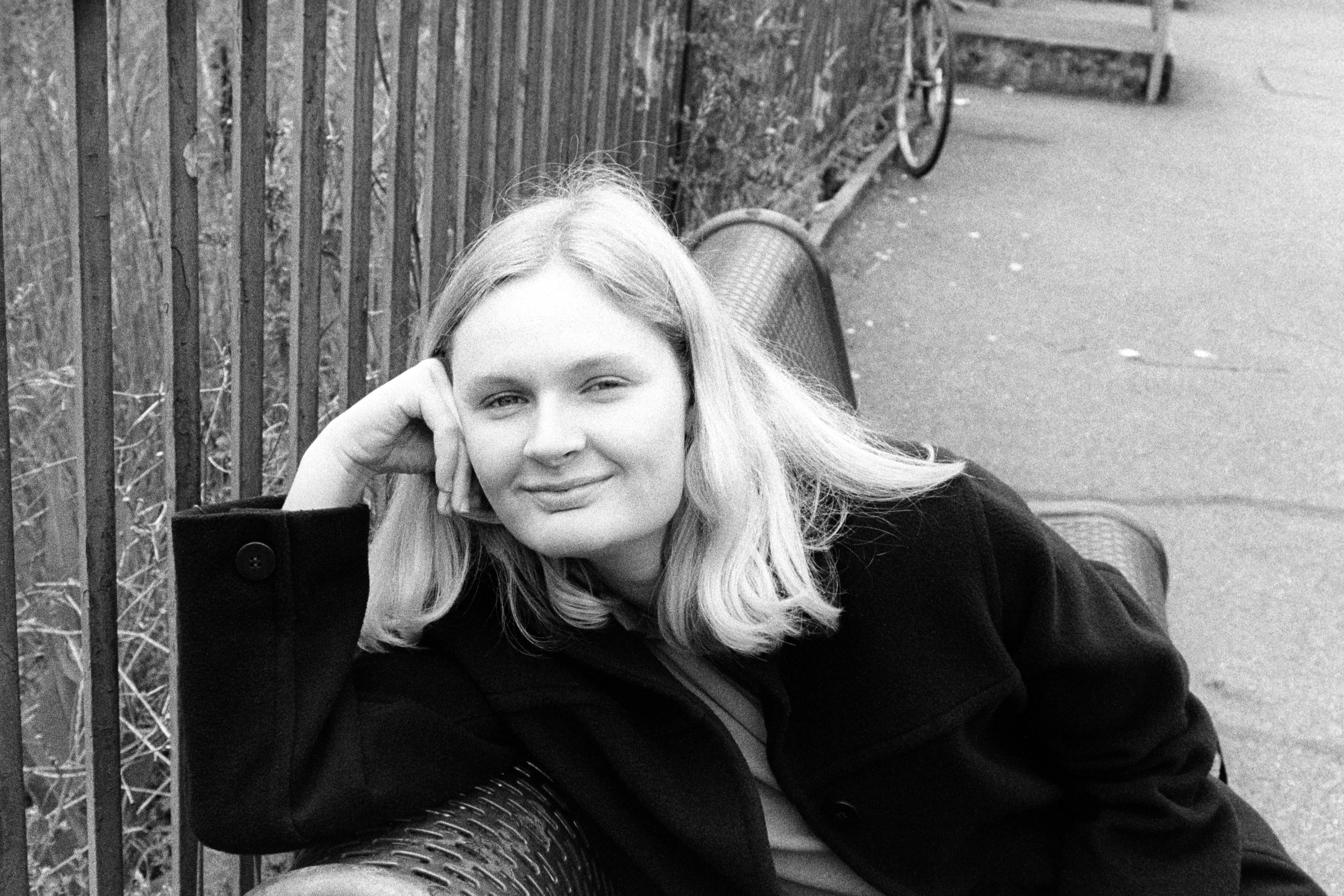 Liddy leaning against a wooden fence on a metal bench, head resting on her hand, smiling softly at the camera.