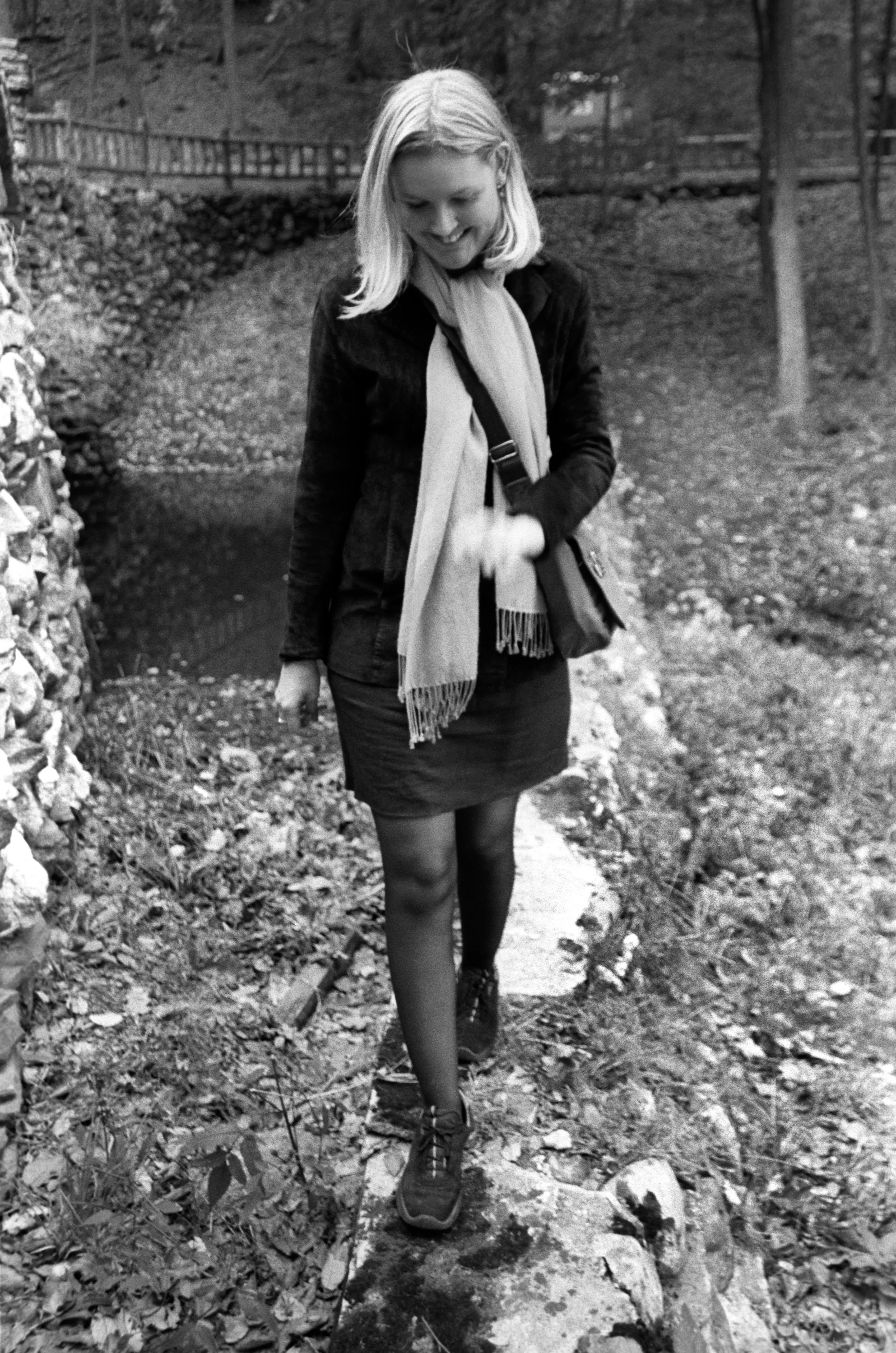Liddy walking along the top of a low stone wall in a wooded park, looking down at her footing, a long scarf draped across her front, smiling softly.