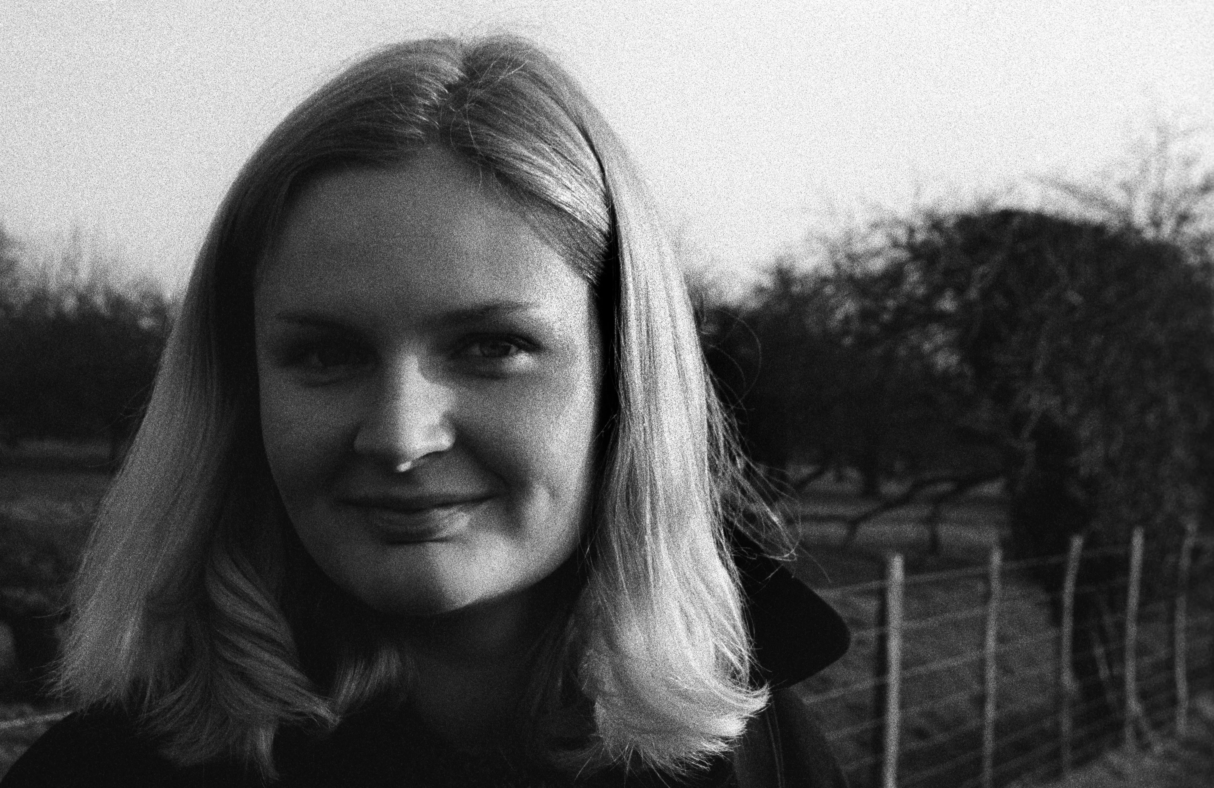 Close portrait of Liddy with a soft closed-mouth smile, dusk light on her hair, a hedgerow and fence-line behind her.