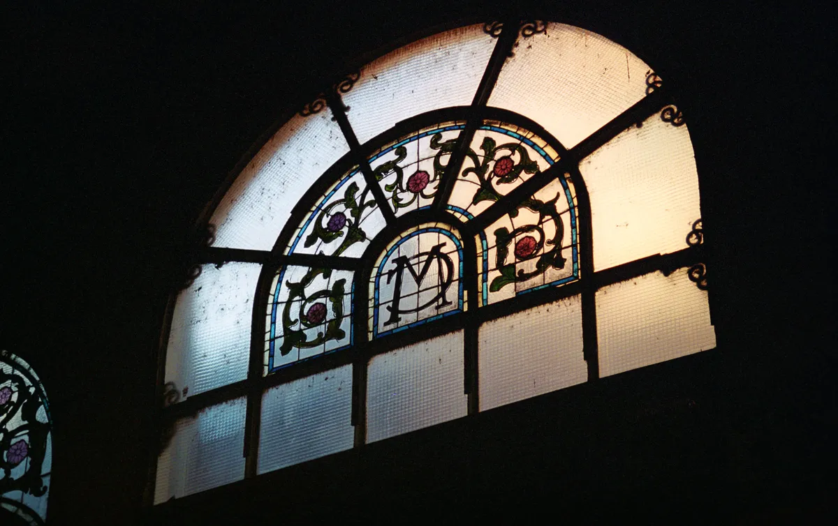An arched market window in the dark, a wrought 'M' at the centre of its stained glass.