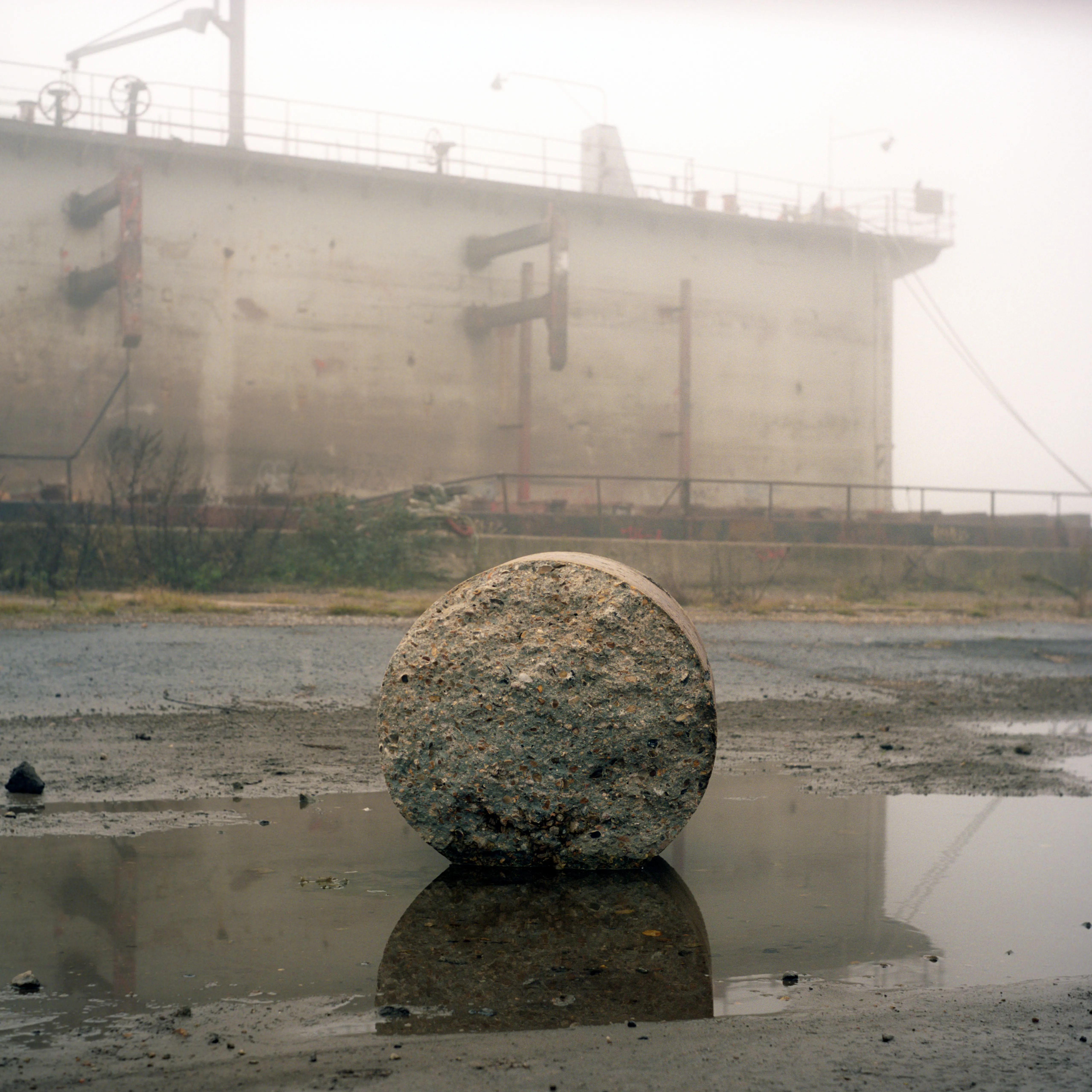 A large spherical buoy left on the muddy tidal foreshore, a rusted hull behind it, reflected in shallow water.