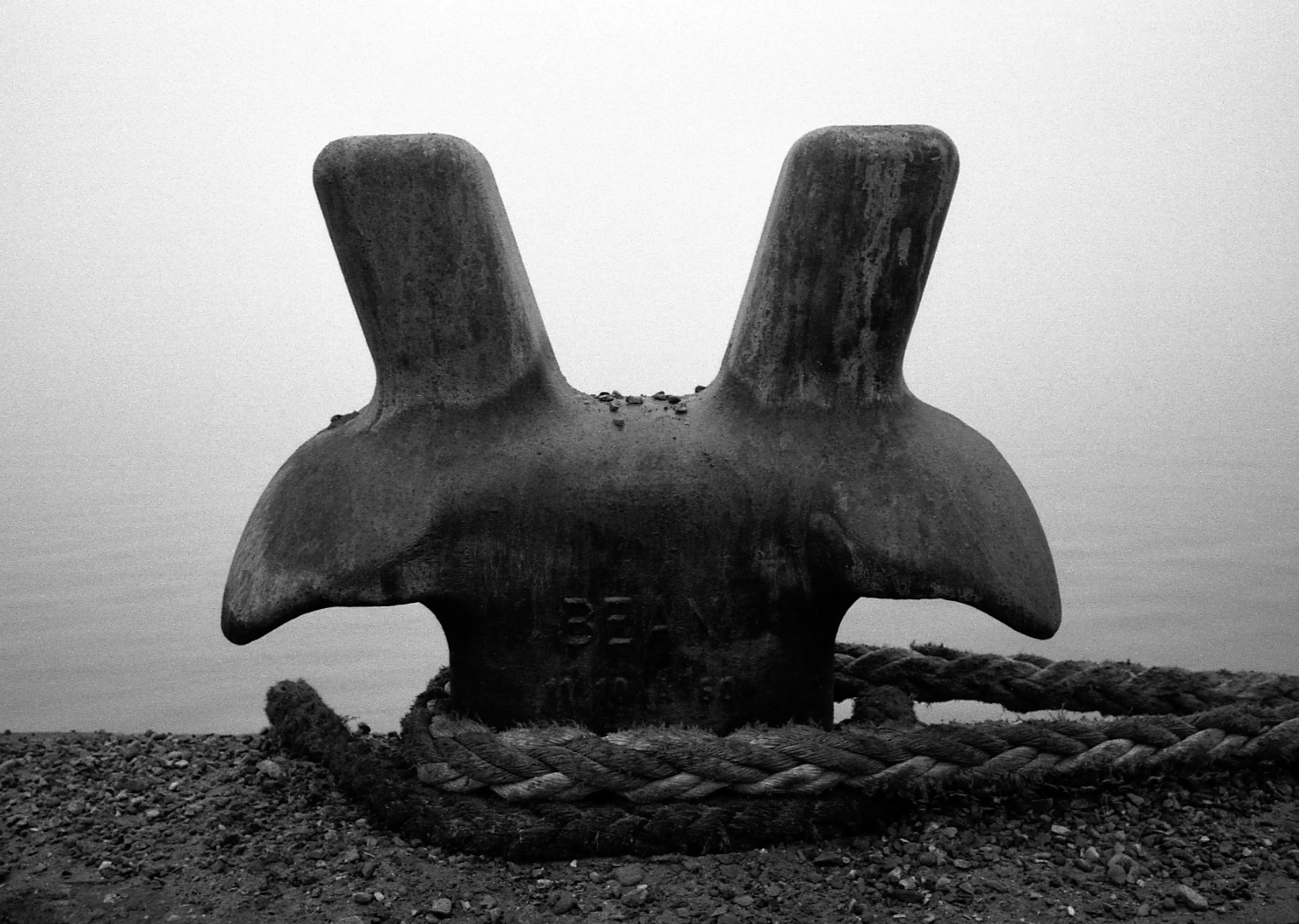 Black-and-white — a heavy mooring bollard with thick knotted rope coiled at its base on a shingle bank by still water.