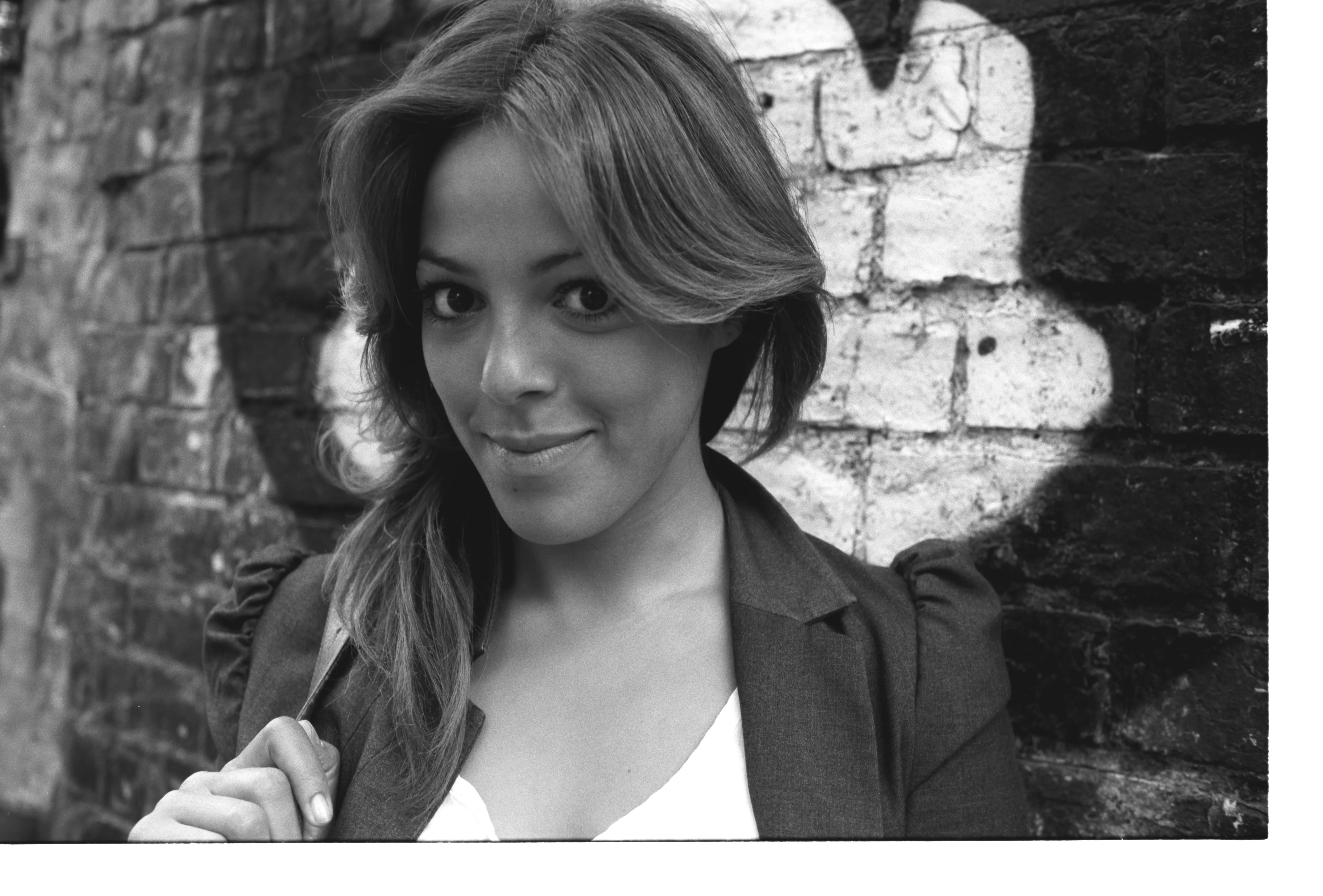 Black-and-white frame: Nawal leans against a graffitied brick wall, looking straight into the lens with a faint, closed-mouth smile.