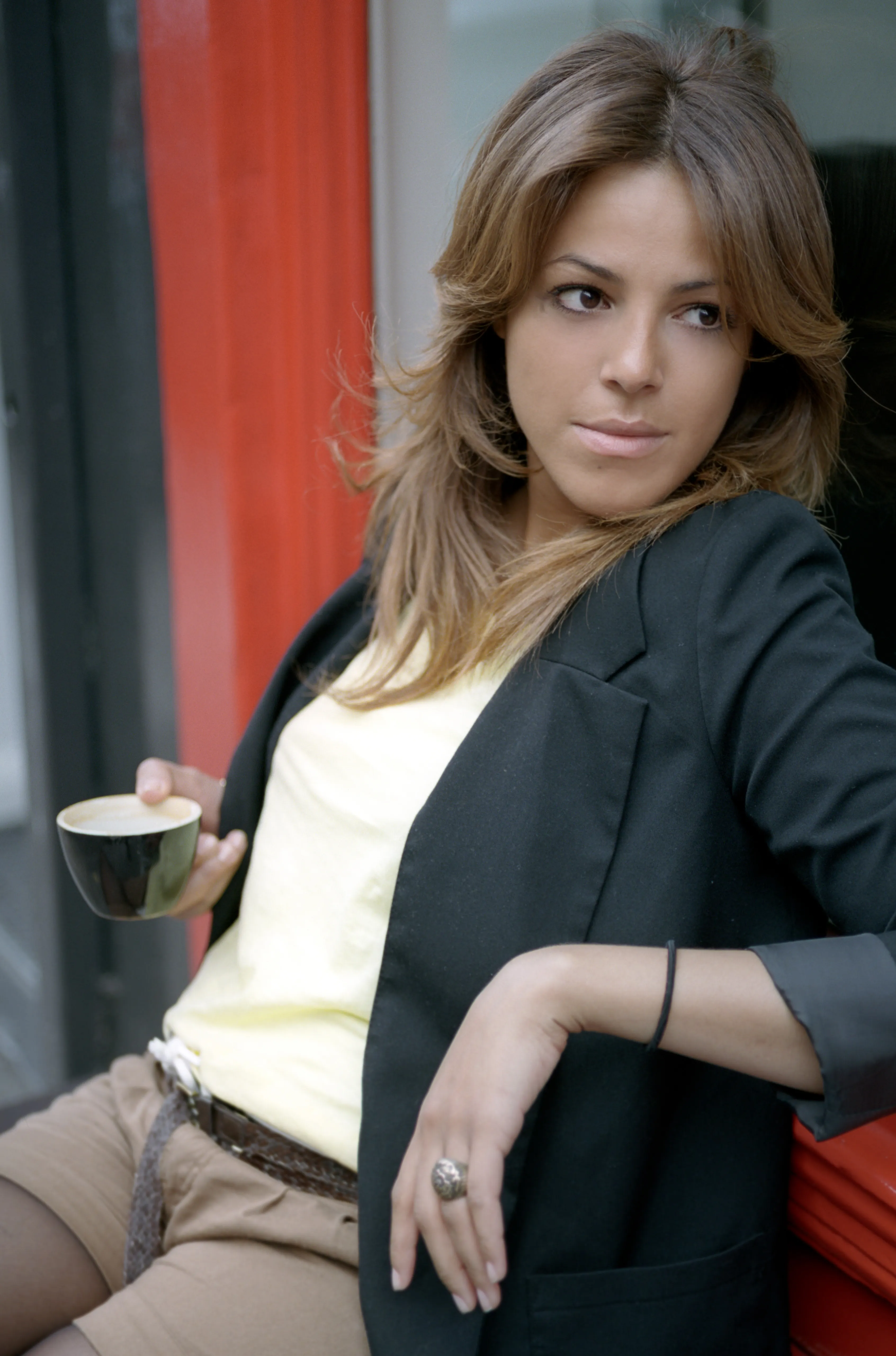 Nawal sits holding a small espresso cup, leaning back against a red shopfront, looking directly at the camera.