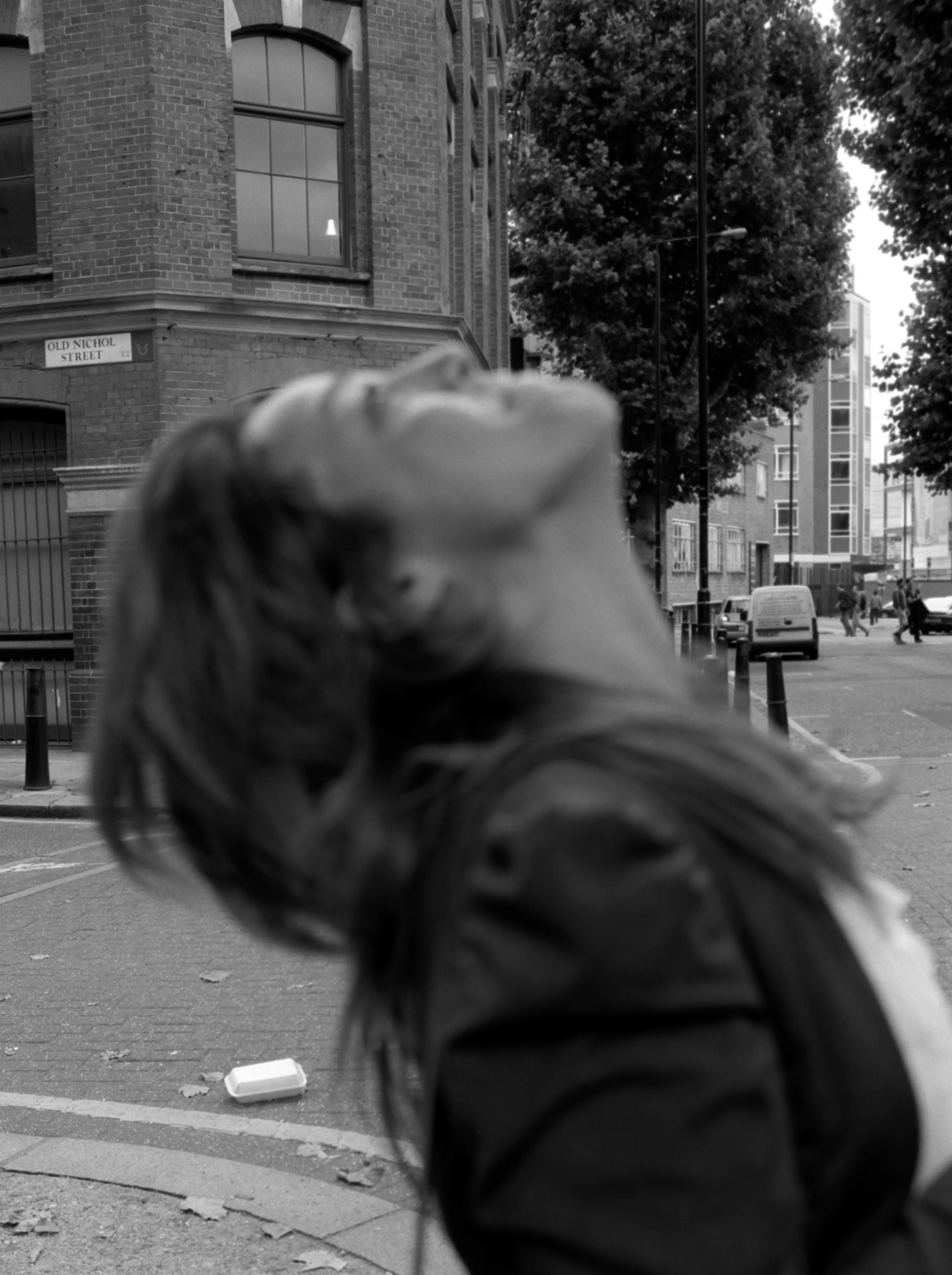 Black-and-white street frame: Nawal tips her head back and laughs, hair swinging, on a quiet London street corner.