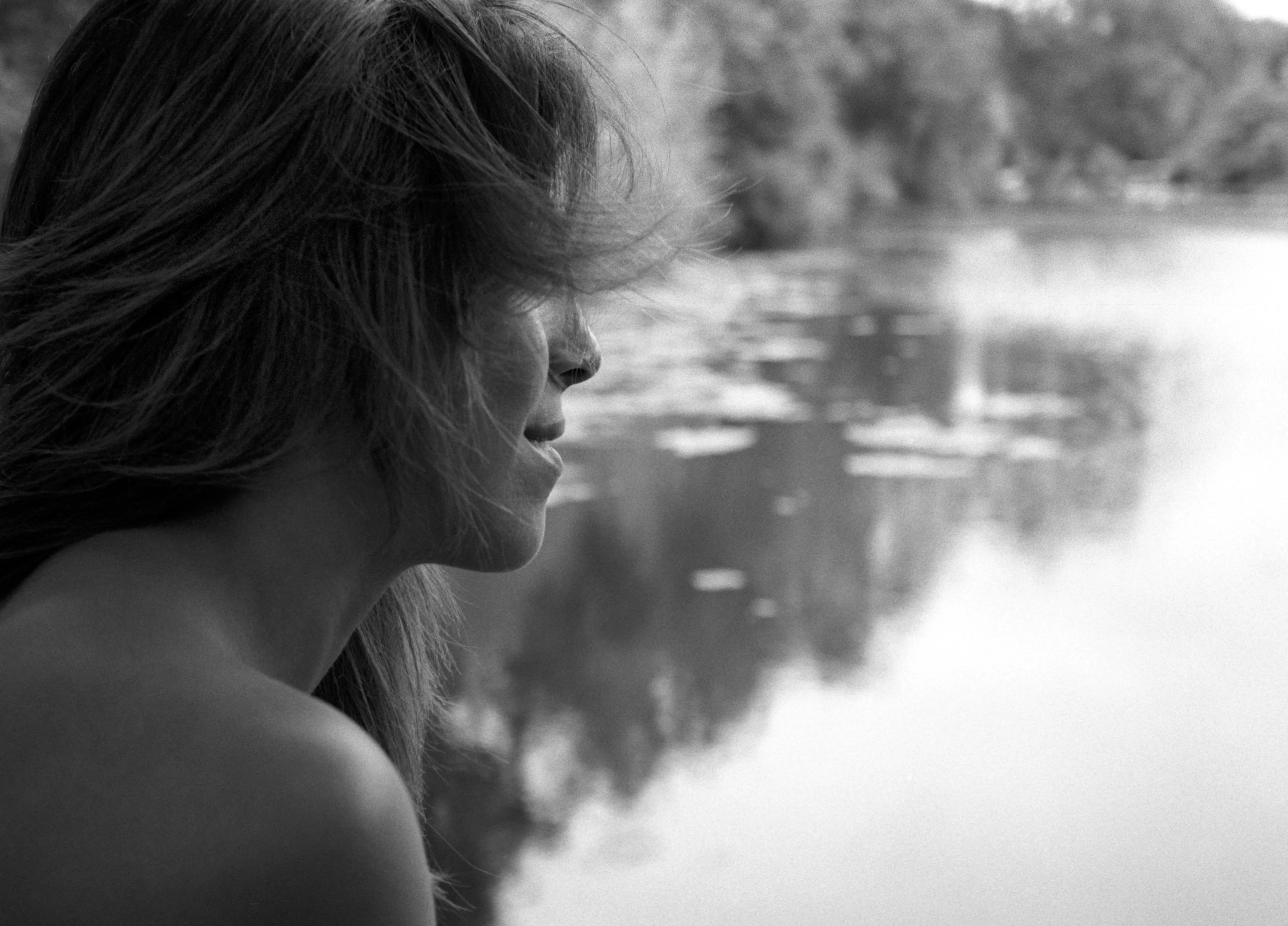 Black-and-white frame: Nawal in profile at the edge of still water, hair across her face, the far bank reflected on the surface.