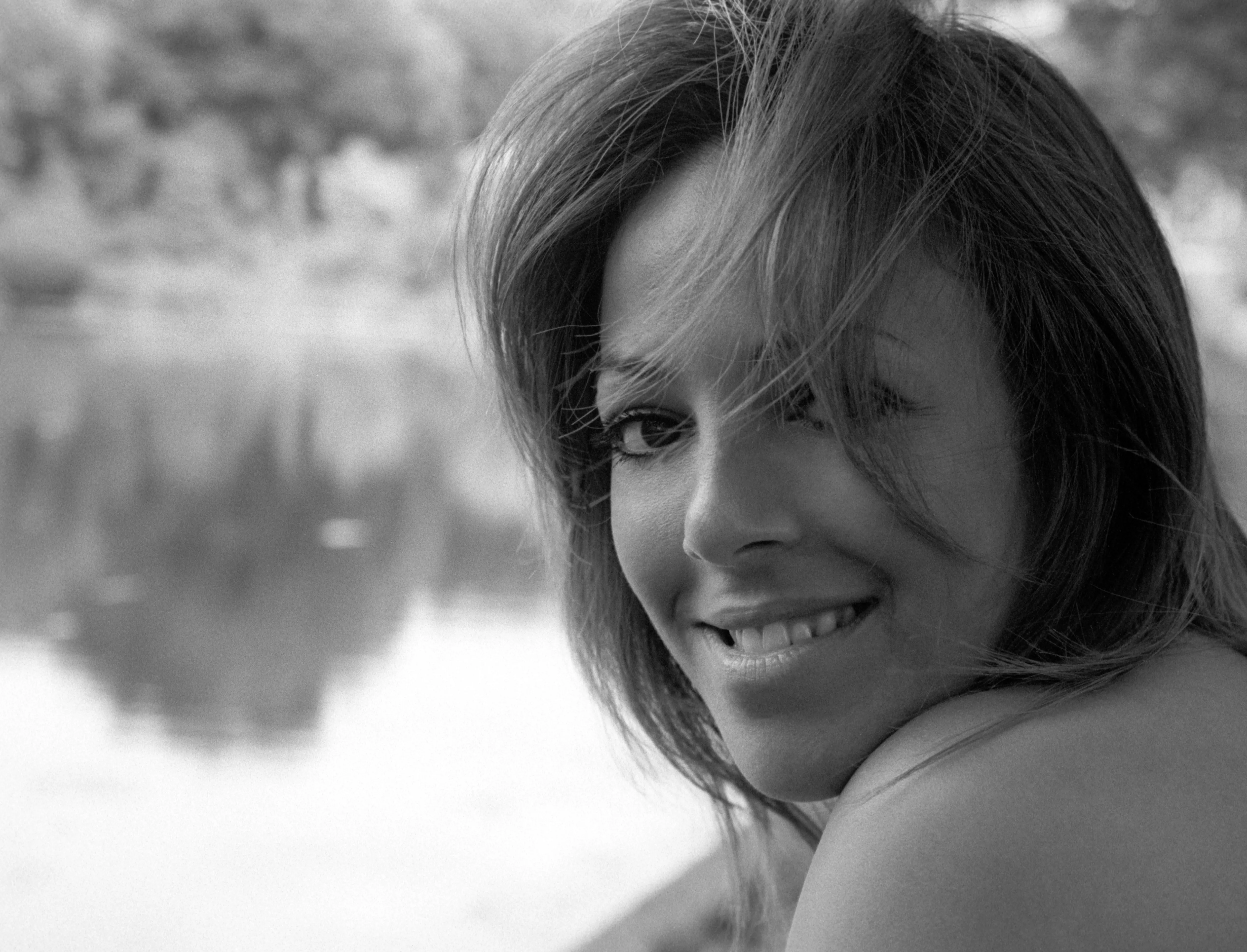 Black-and-white frame: Nawal turns over her shoulder with a broad, easy smile, trees and water blurred behind her.