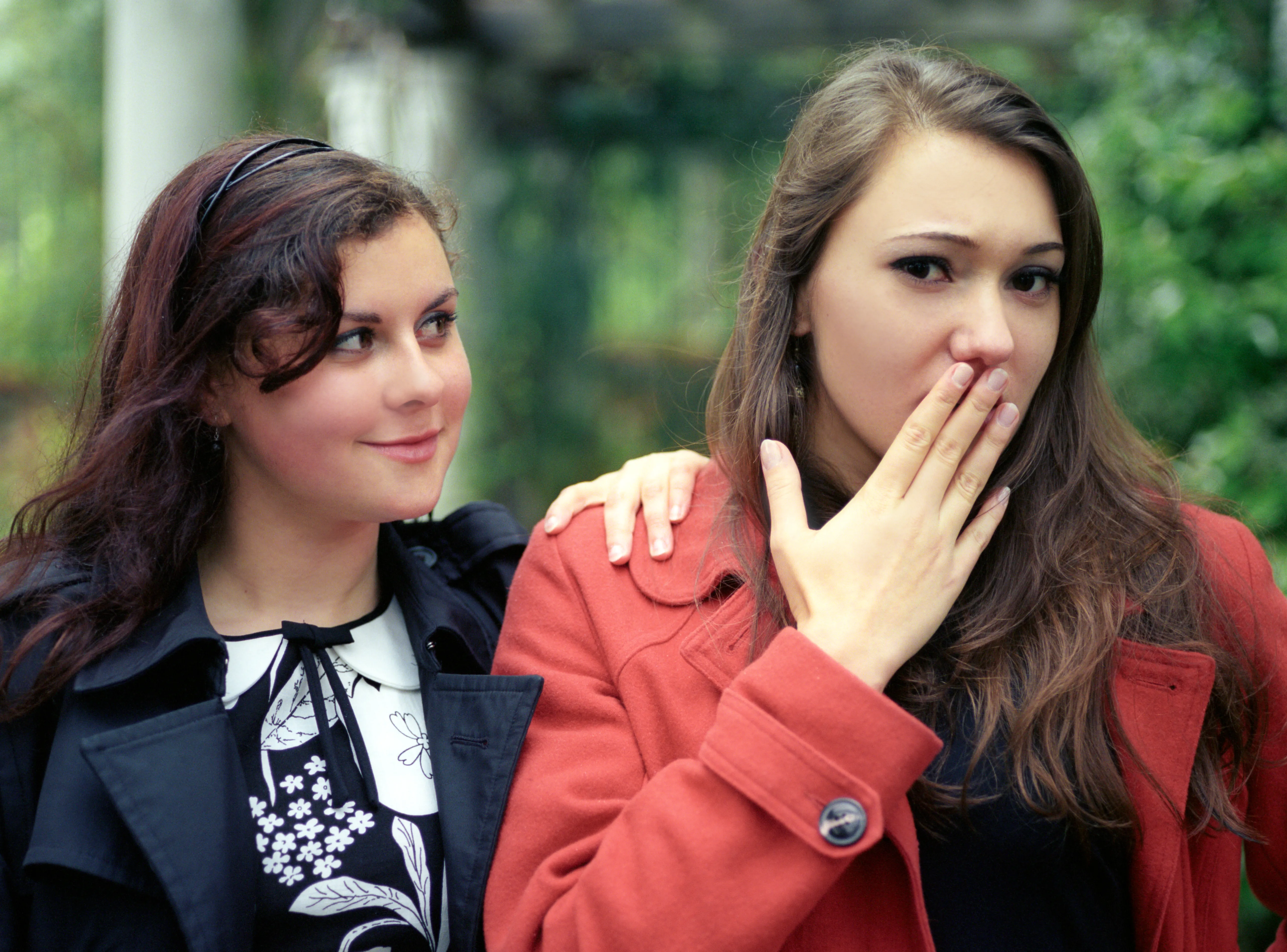 Two young women outdoors; one in a red coat raises a hand to her mouth while the other, in a dark coat, looks towards her.