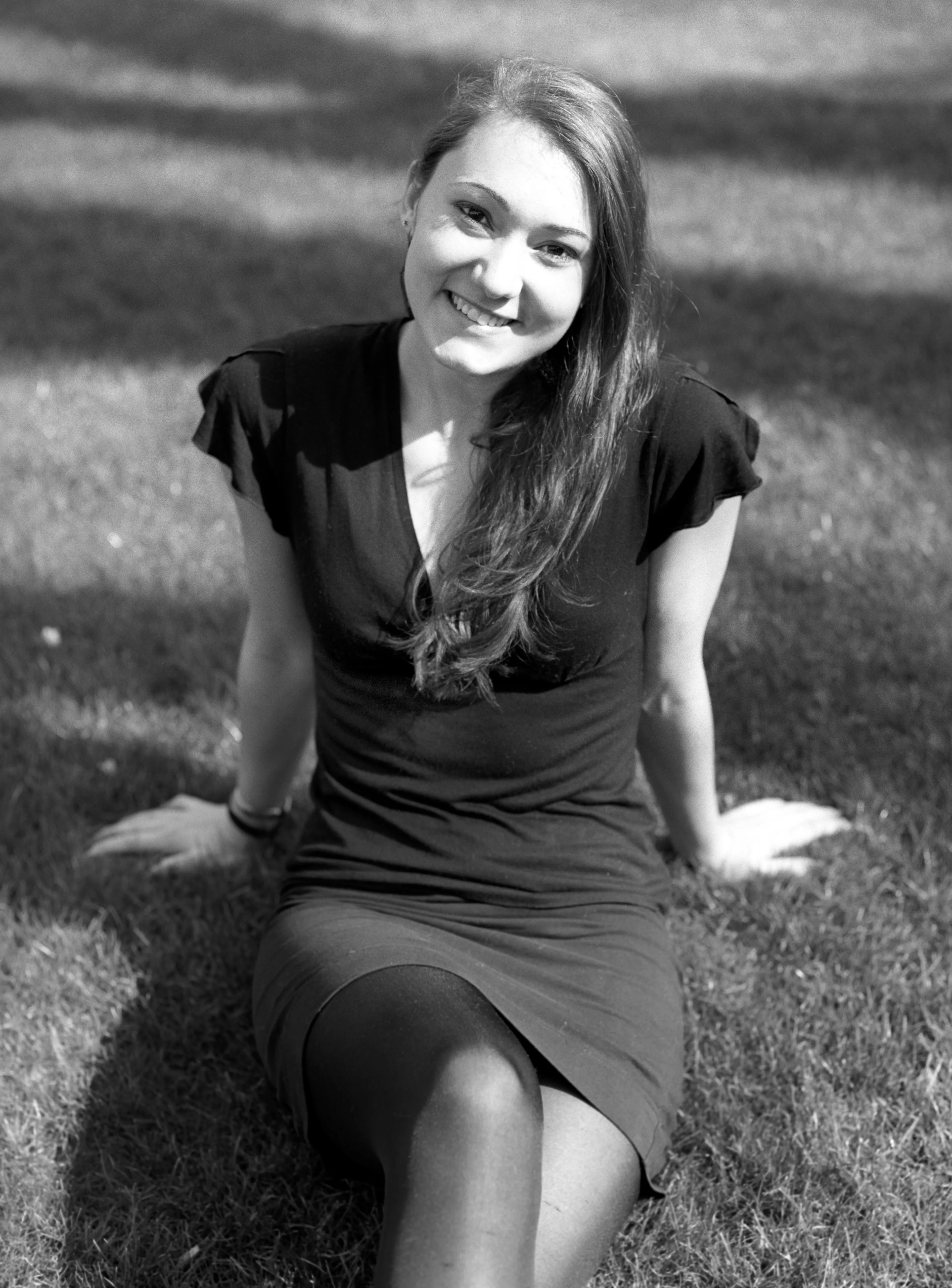 Black-and-white photograph of a young woman in a dark dress sitting on grass, smiling at the camera.