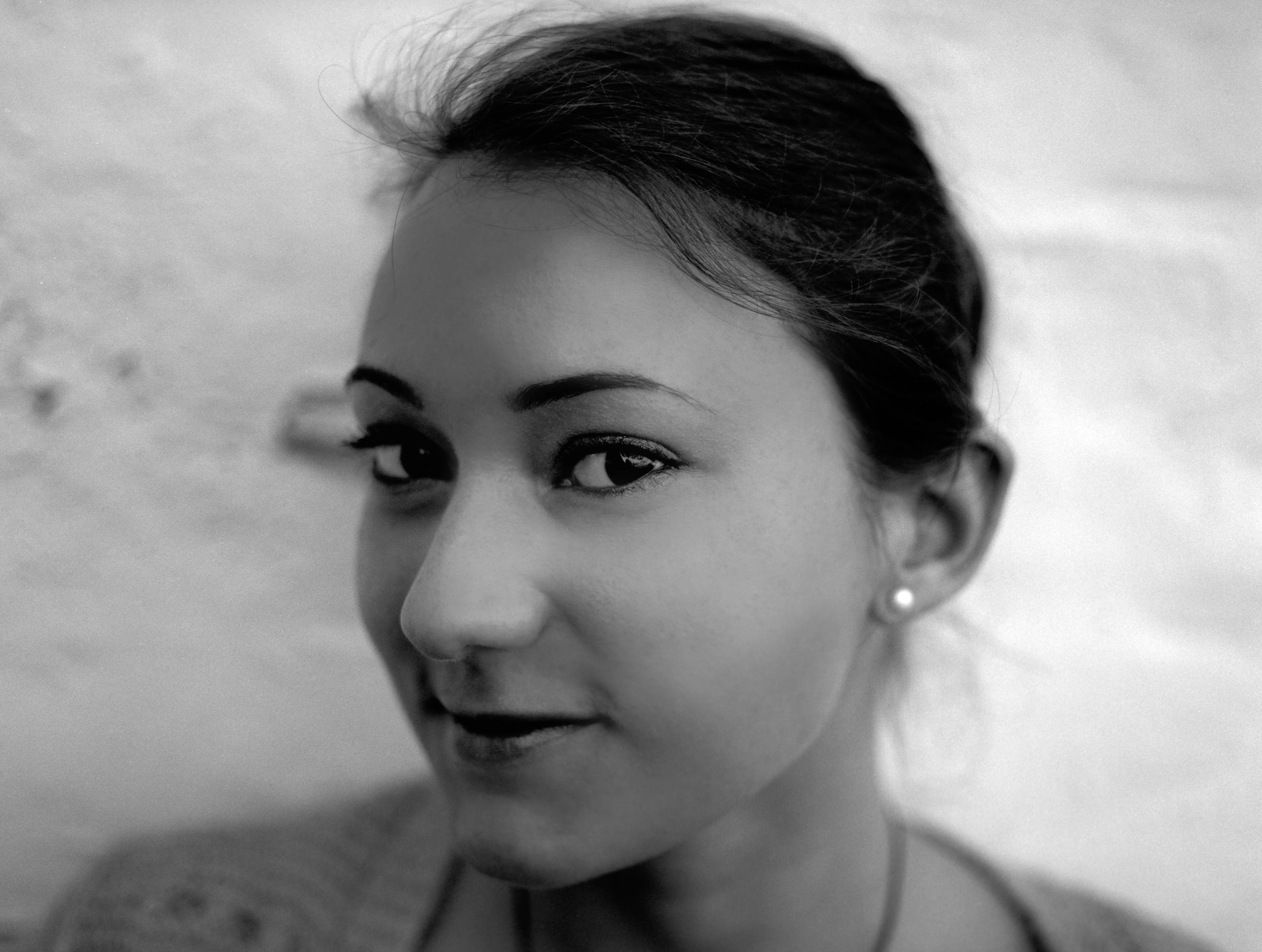 Close black-and-white portrait of a young woman, hair tied back, turned slightly towards the camera against a pale plaster wall.