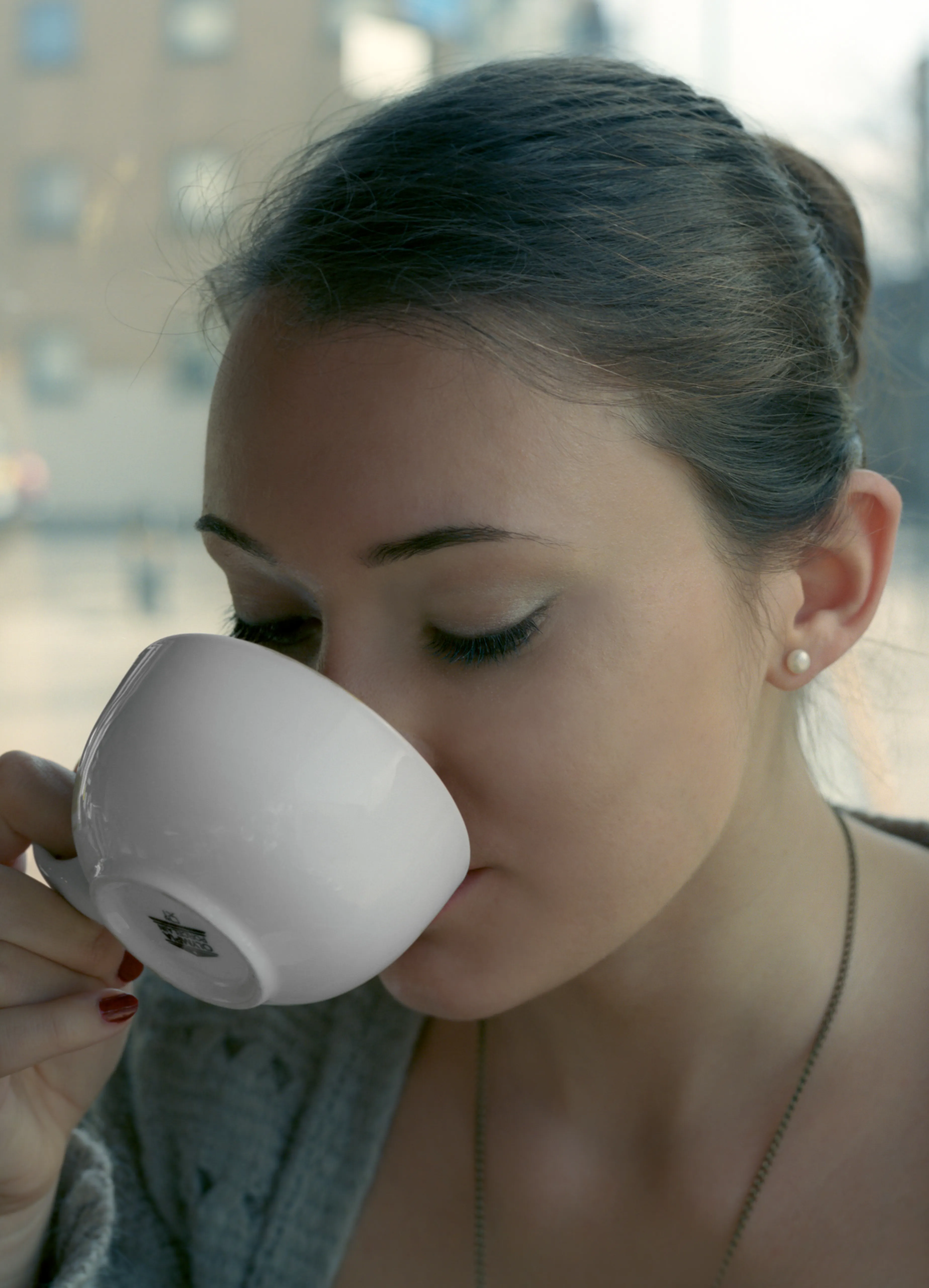 A young woman drinking from a white cup, eyes closed, hair tied back, in soft daylight.