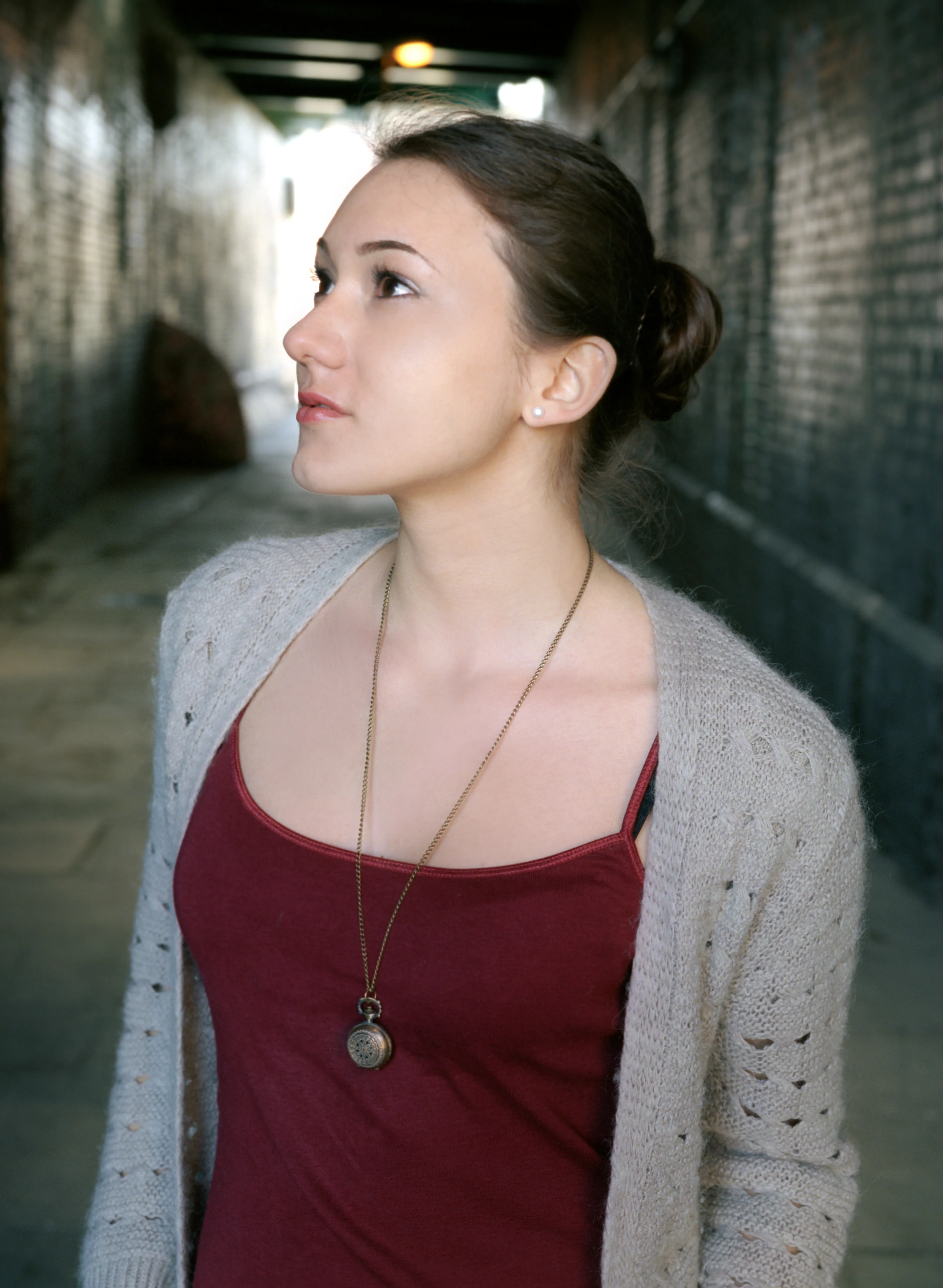 A young woman in a red top and pale cardigan standing in a brick archway, looking upward towards the light.