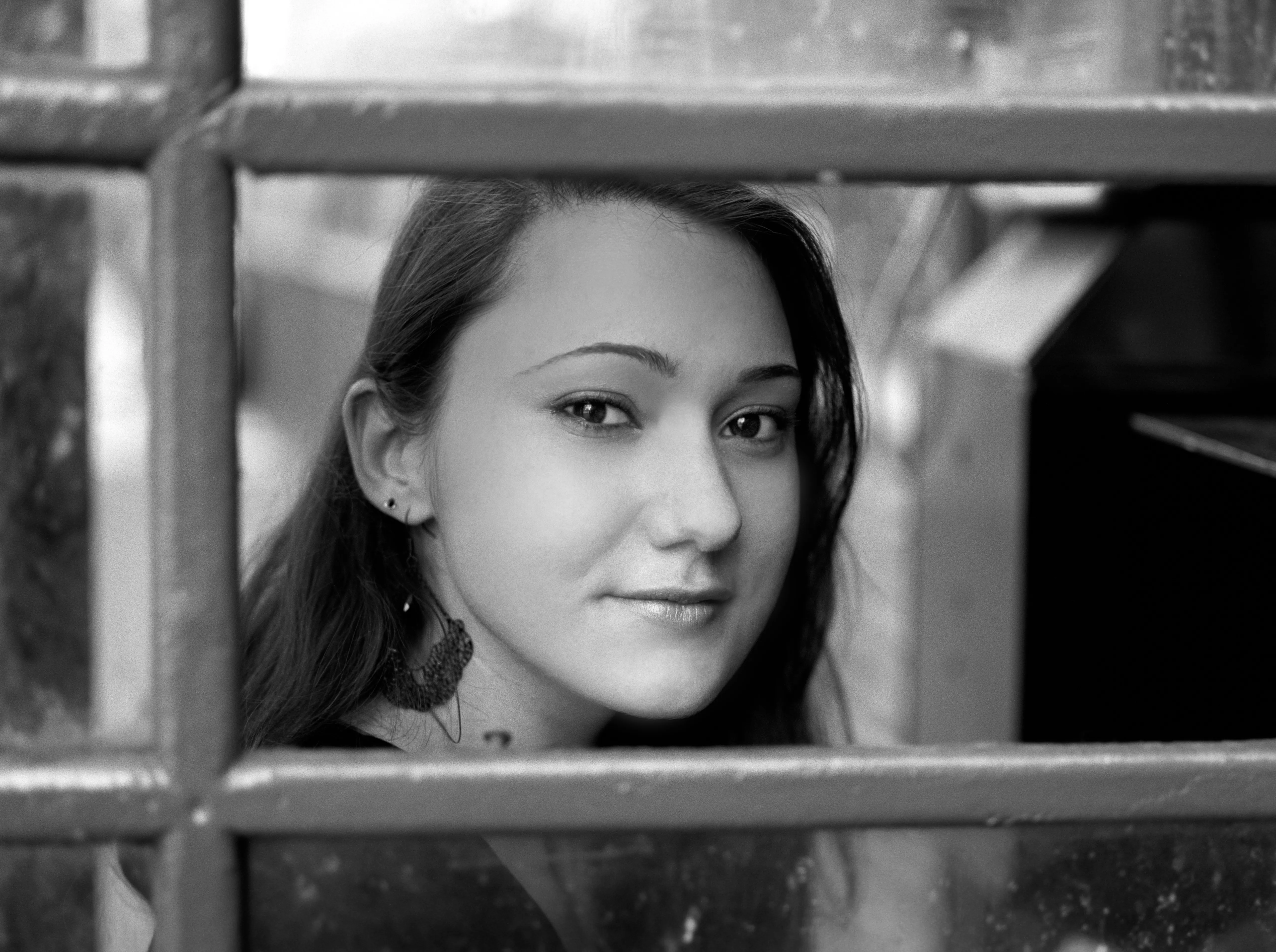 Black-and-white portrait of a young woman framed behind horizontal metal railings, looking at the camera.