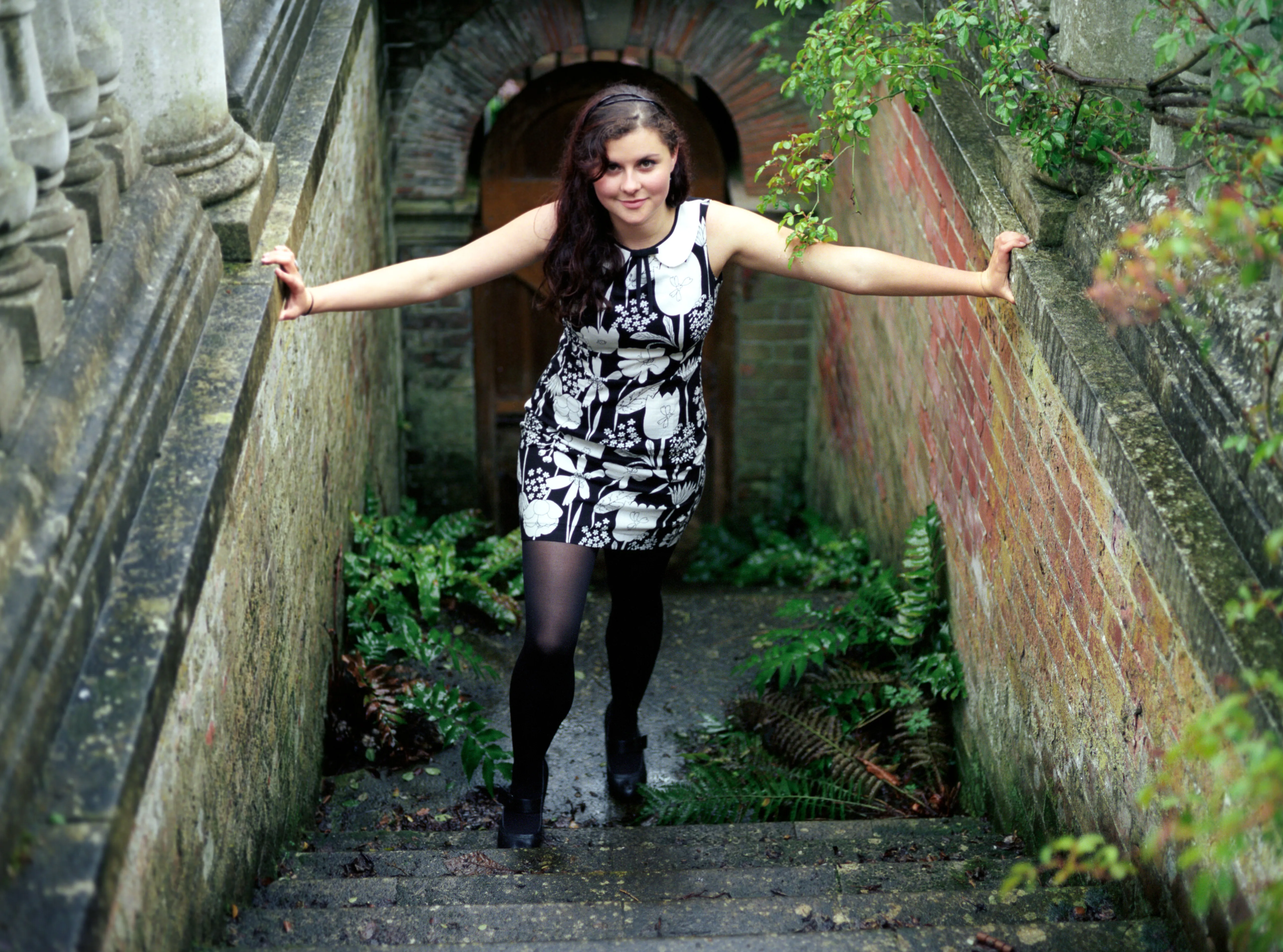A young woman in a patterned dress standing in a sunken stone passage, arms outstretched to the walls on either side.