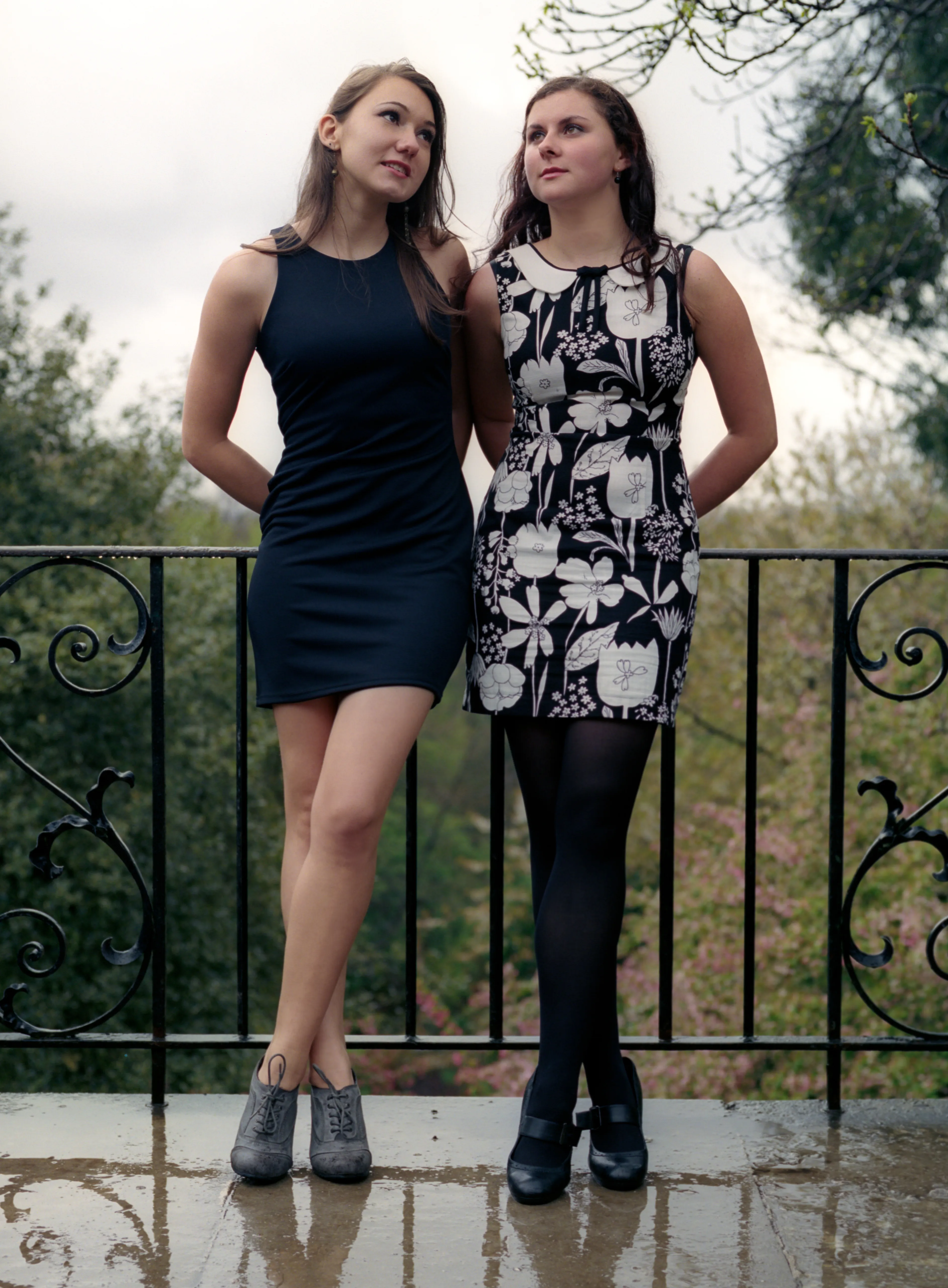 Two young women in dresses standing at a wrought-iron railing, looking upward, a wet stone terrace beneath them.