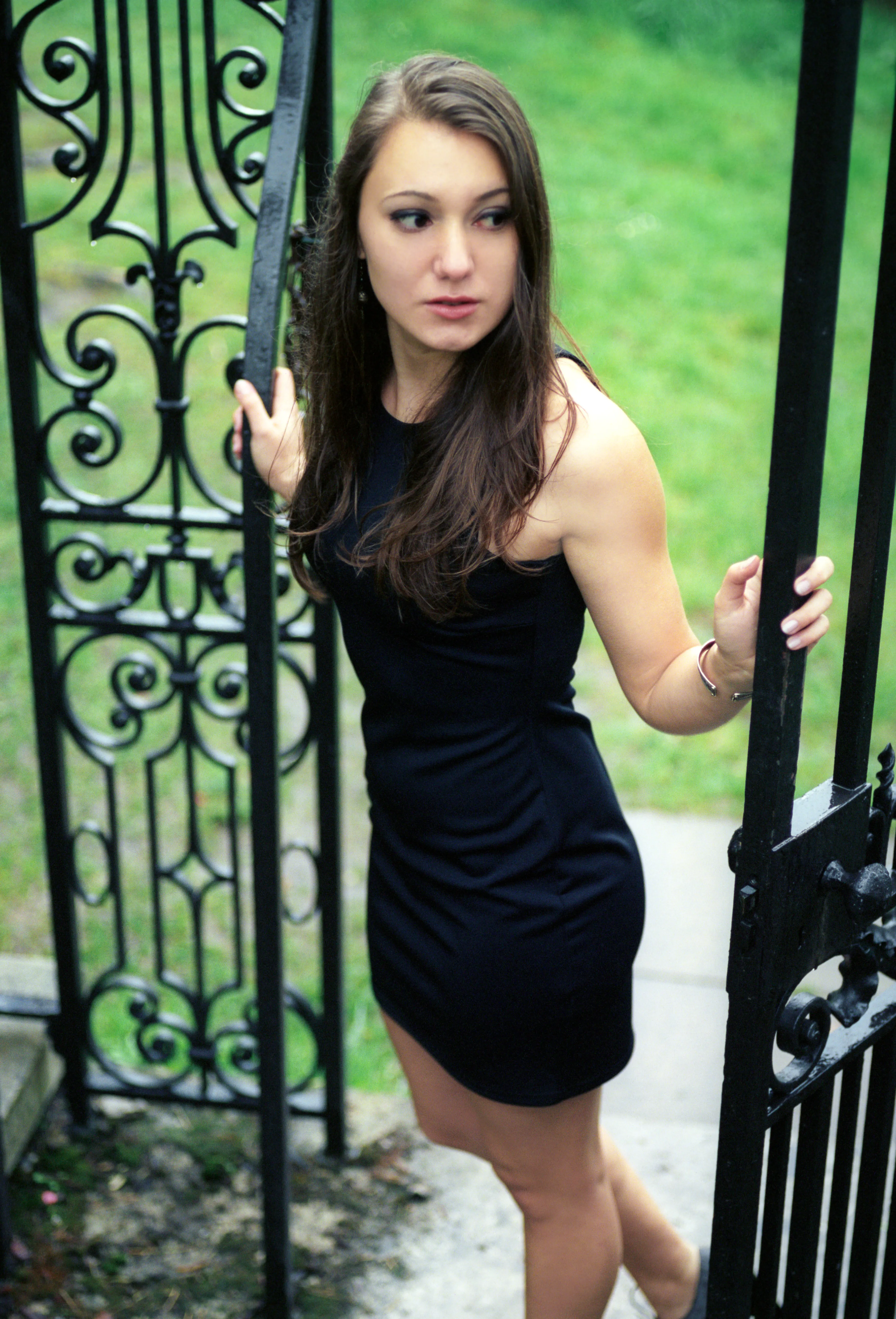 A young woman in a black dress standing in an open wrought-iron gate, one hand on the bars, grass behind.