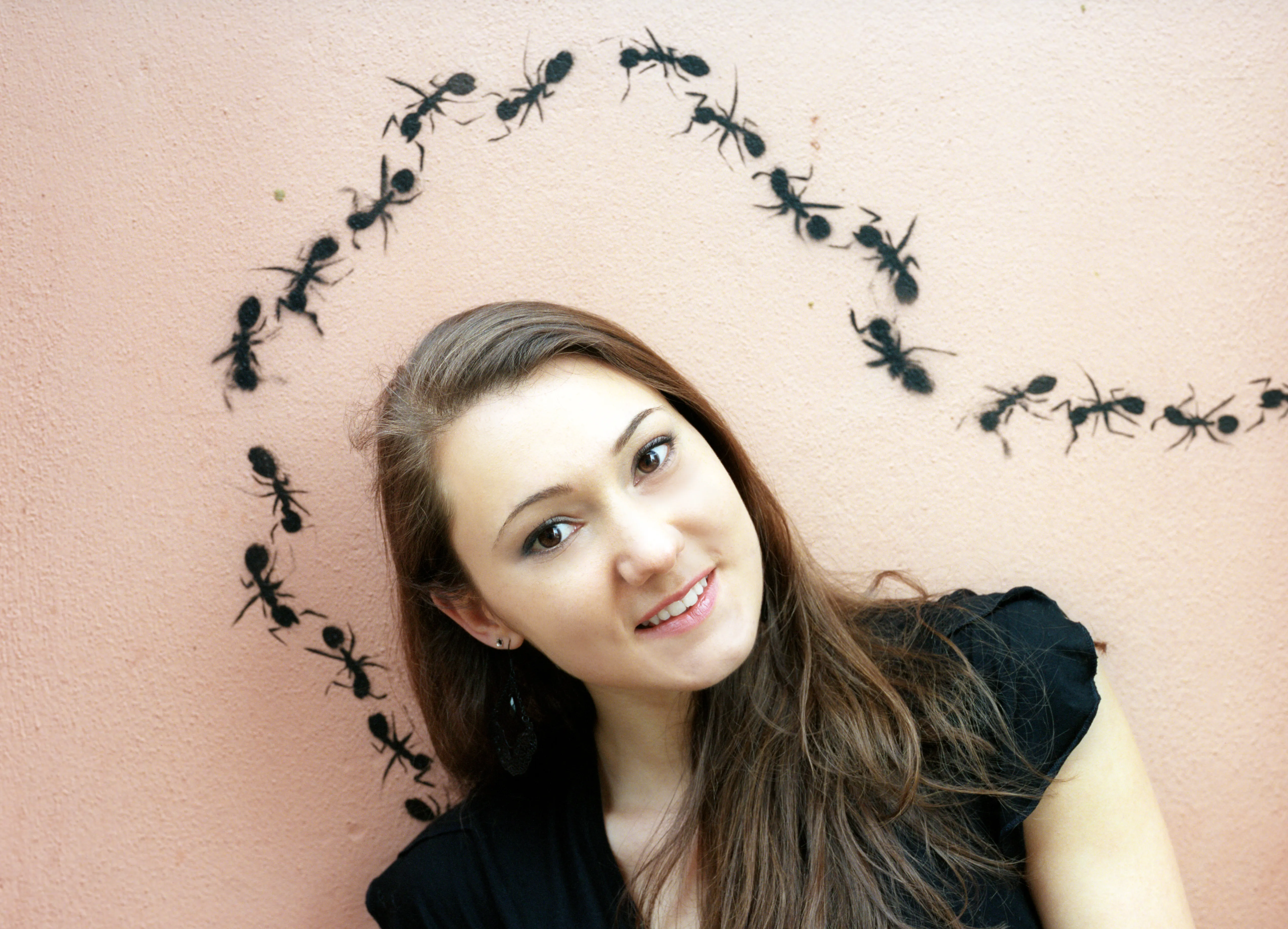 A young woman beside a pink wall on which black ants are arranged in a curving arc above her head.