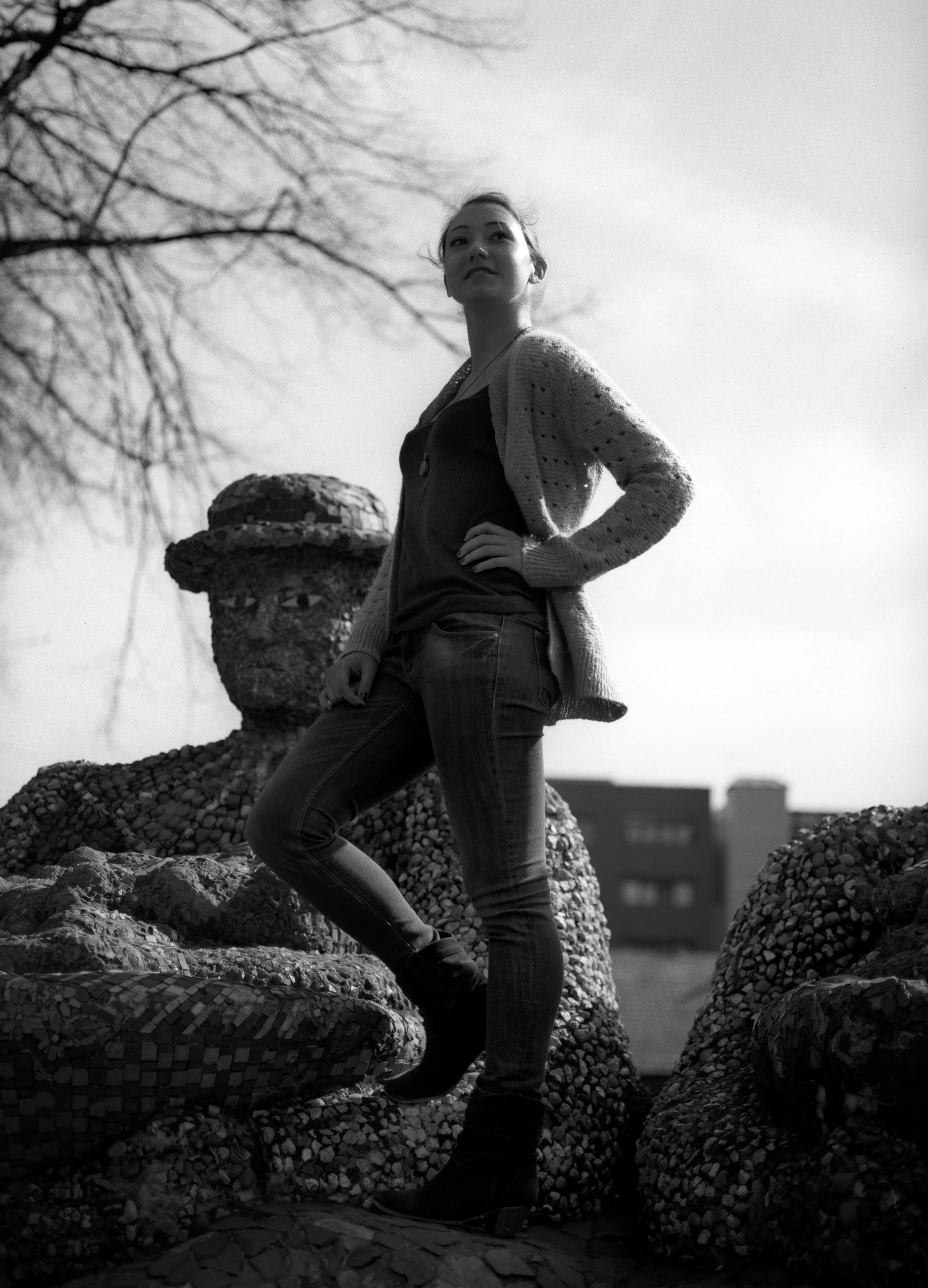 Black-and-white photograph of a figure standing on a stone garden ornament against a bright sky, one hand on the hip.