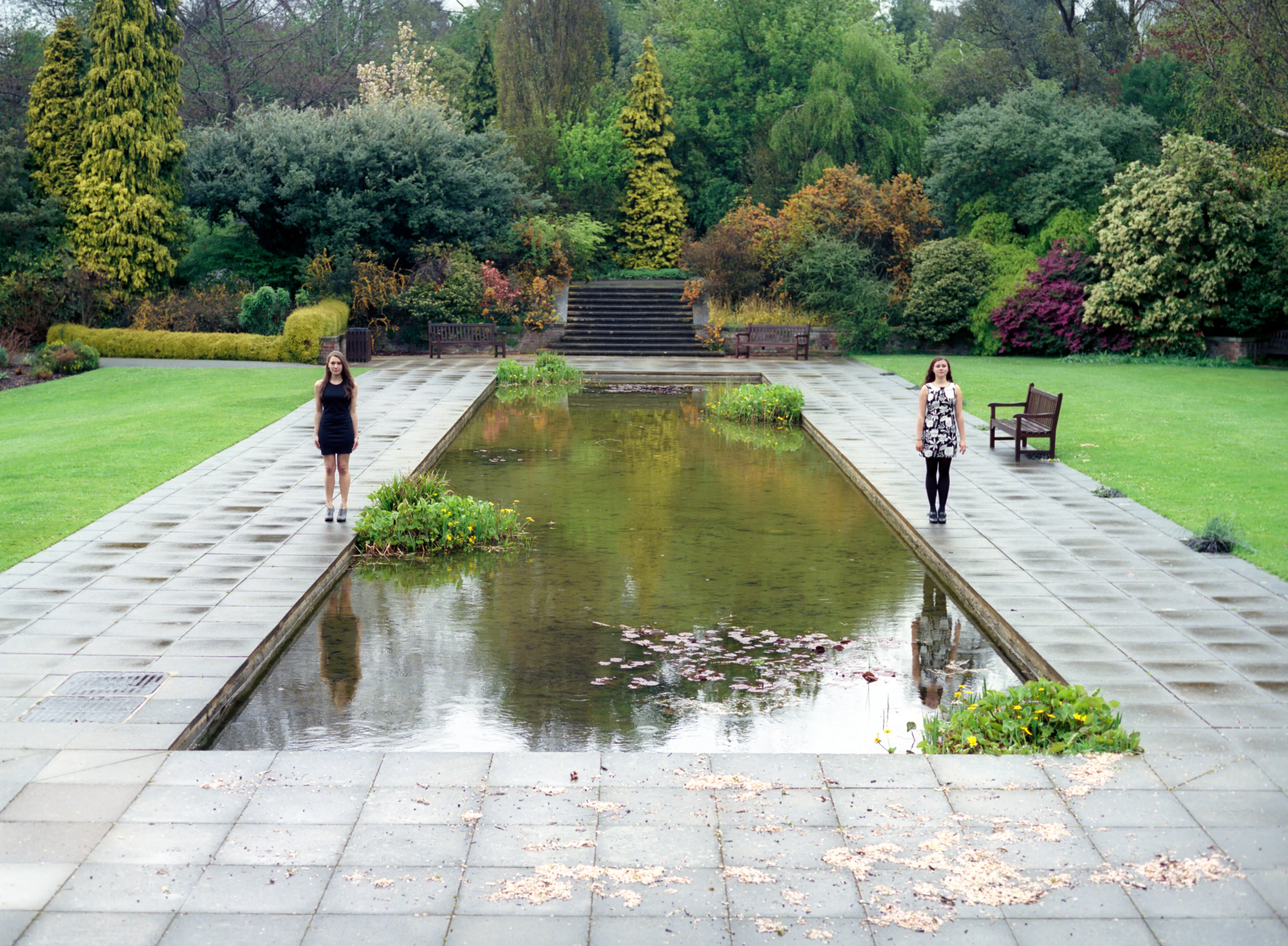 Two distant figures standing at either end of a long rectangular reflecting pool in a formal garden.