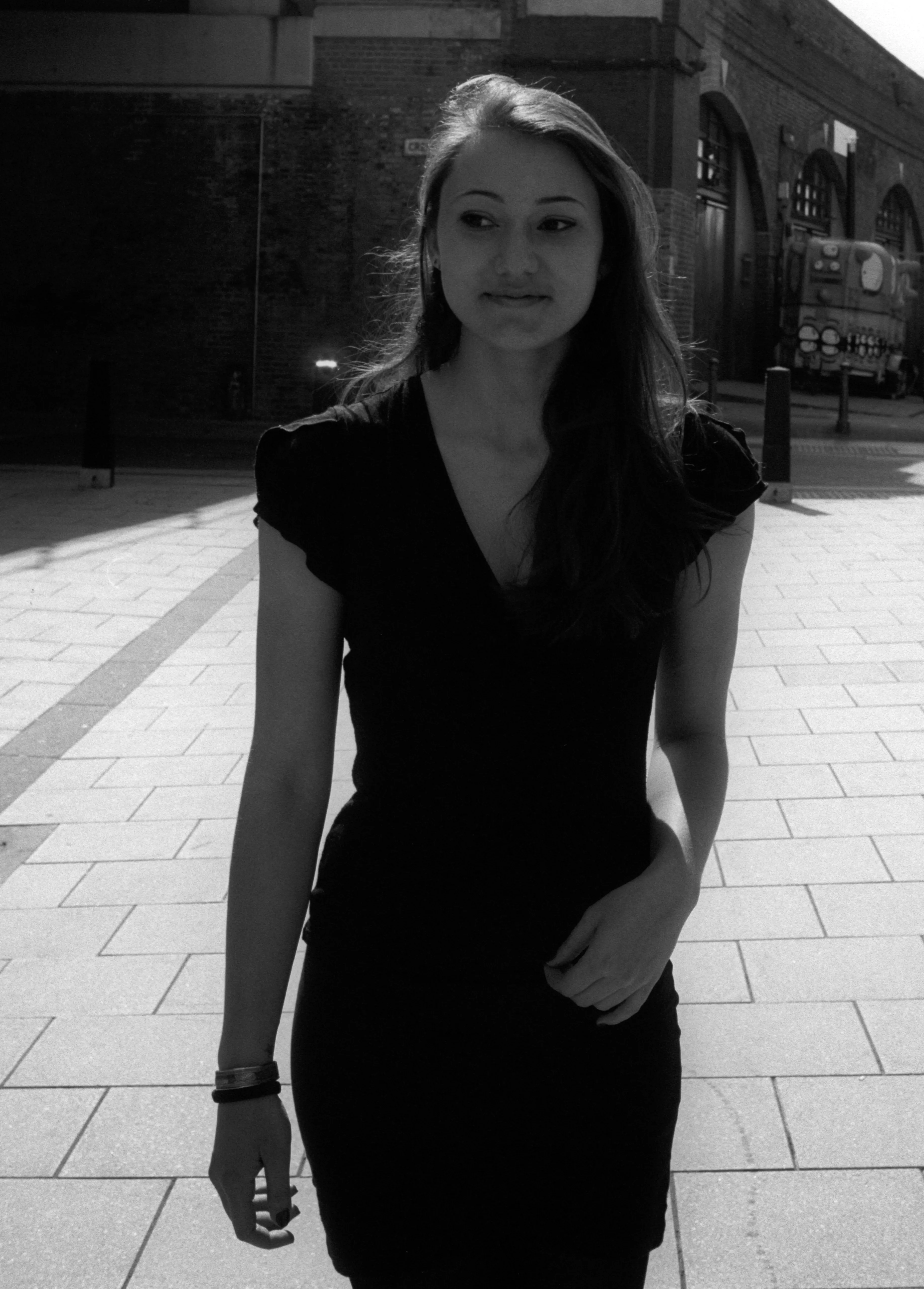 Black-and-white photograph of a young woman in a dark dress walking along a paved street.