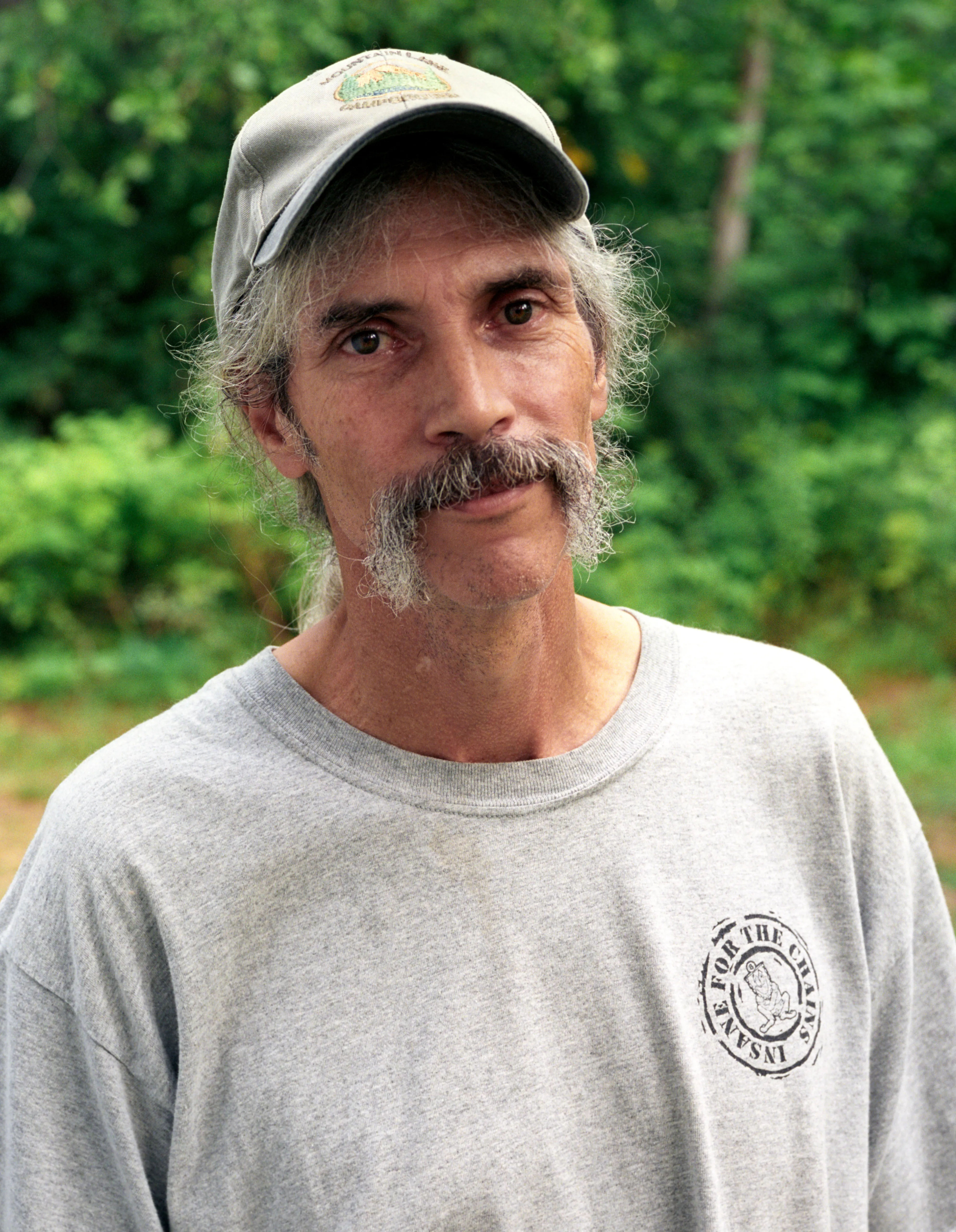 The man outside his house that morning — long grey hair under a cap, a moustache, summer green behind him.