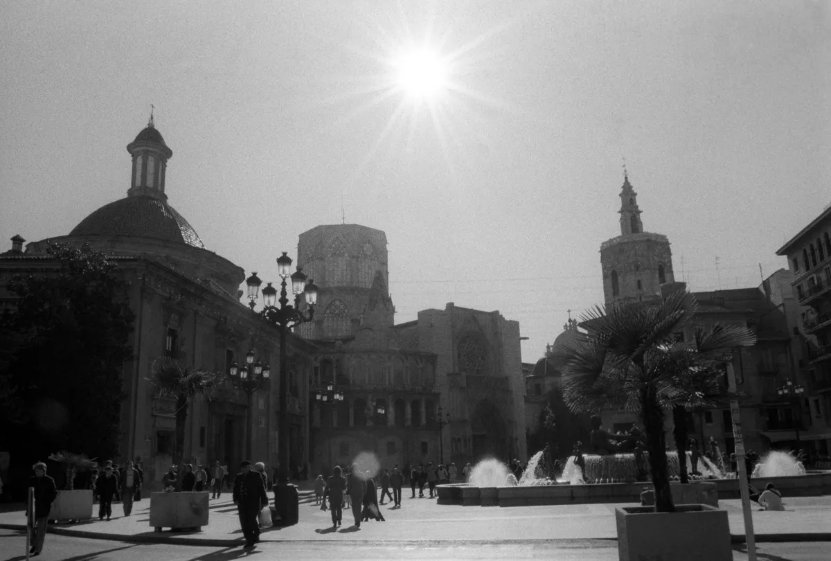 Sun bursting over the Plaza de la Virgen — basilica, cathedral and the Miguelete, people crossing with the fountain behind.