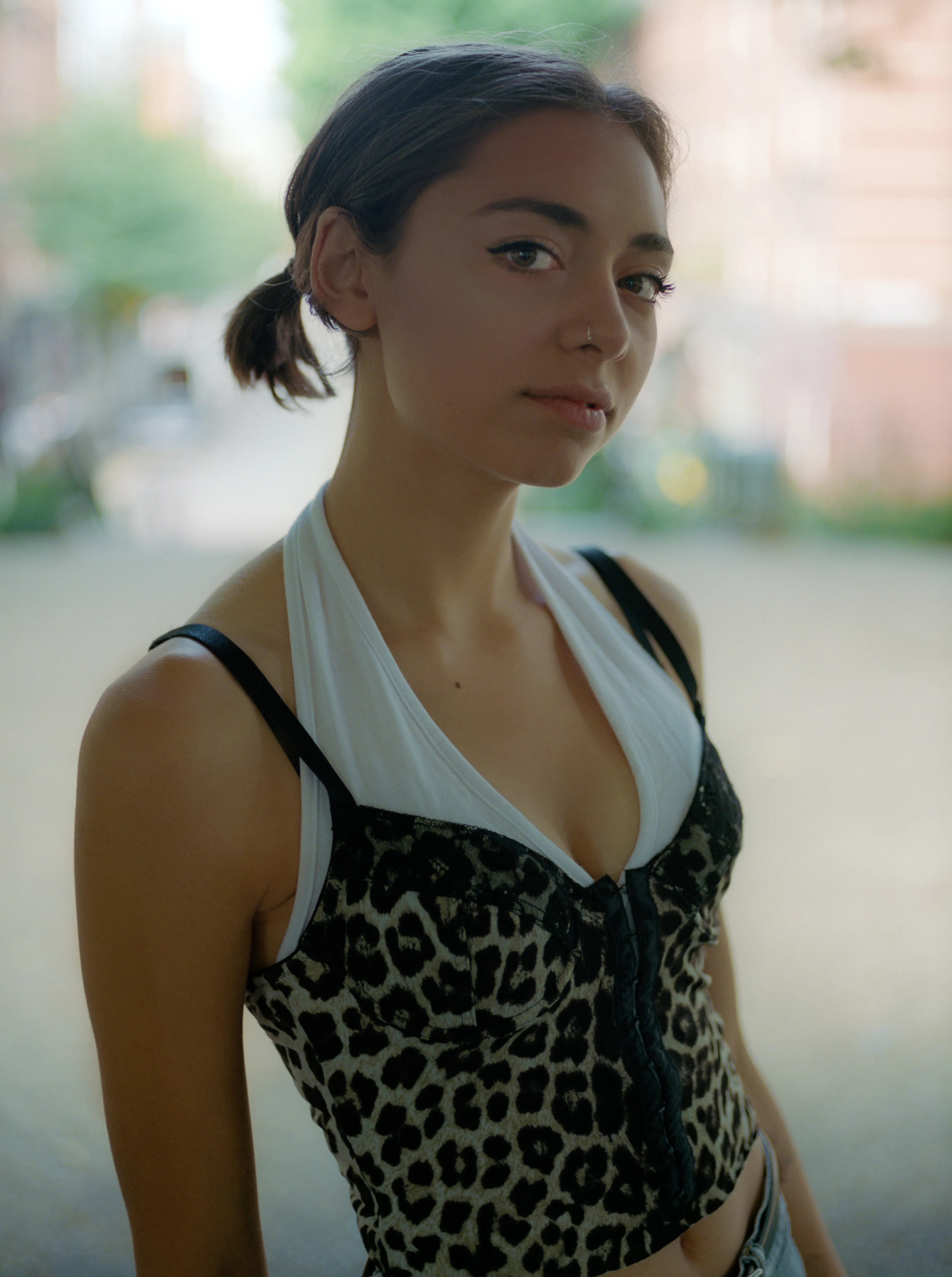 Riya stands on a city pavement in a leopard-print corset over a white halter top, facing the camera, the street behind her out of focus.