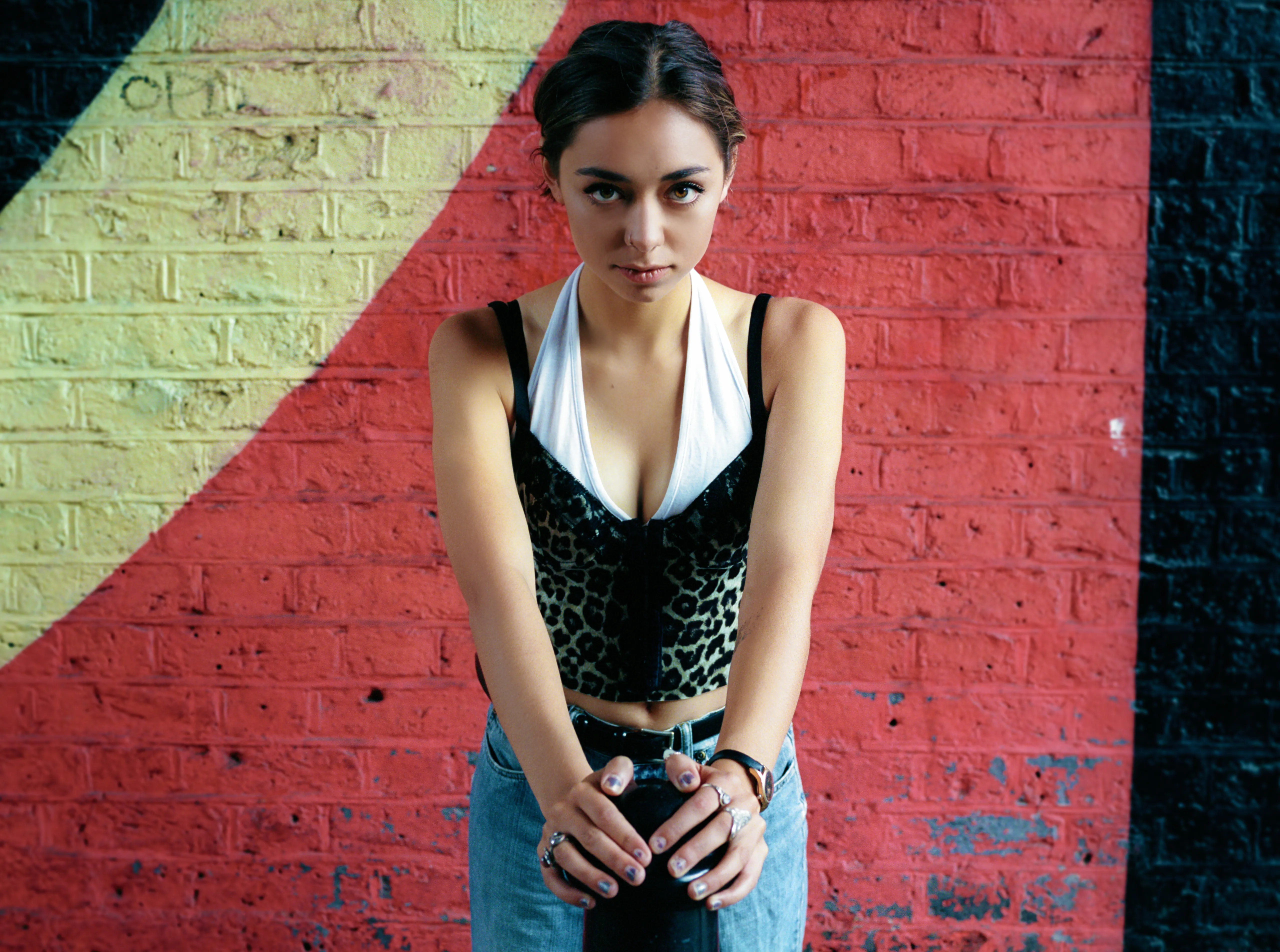 Riya leans forward with her hands clasped low, looking intently at the camera, against a brick wall painted in red, yellow and black.