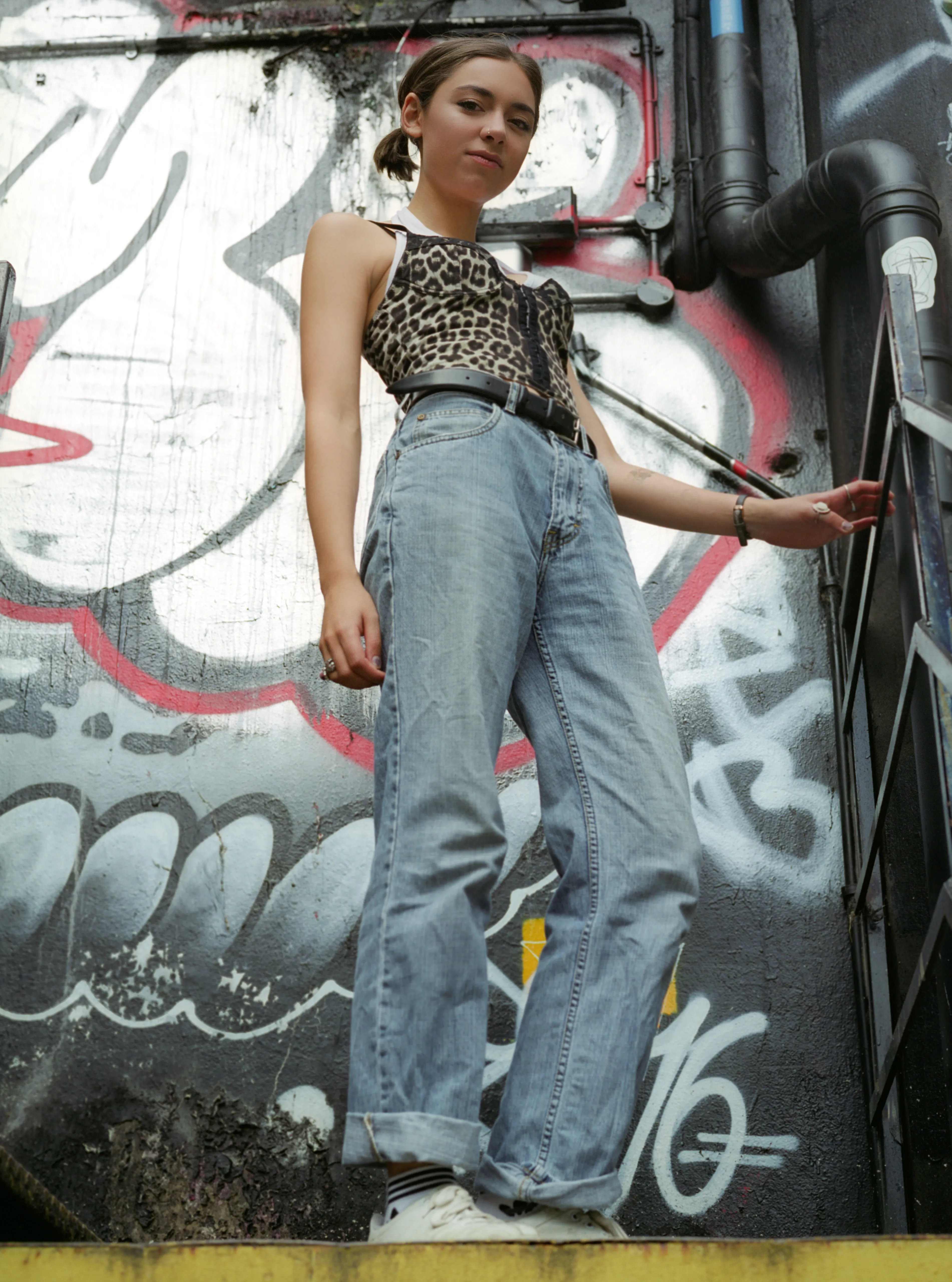 A low-angle, full-length view of Riya standing on a stairway in a leopard-print top and loose jeans, one hand on a metal rail, a graffiti-covered wall behind her.