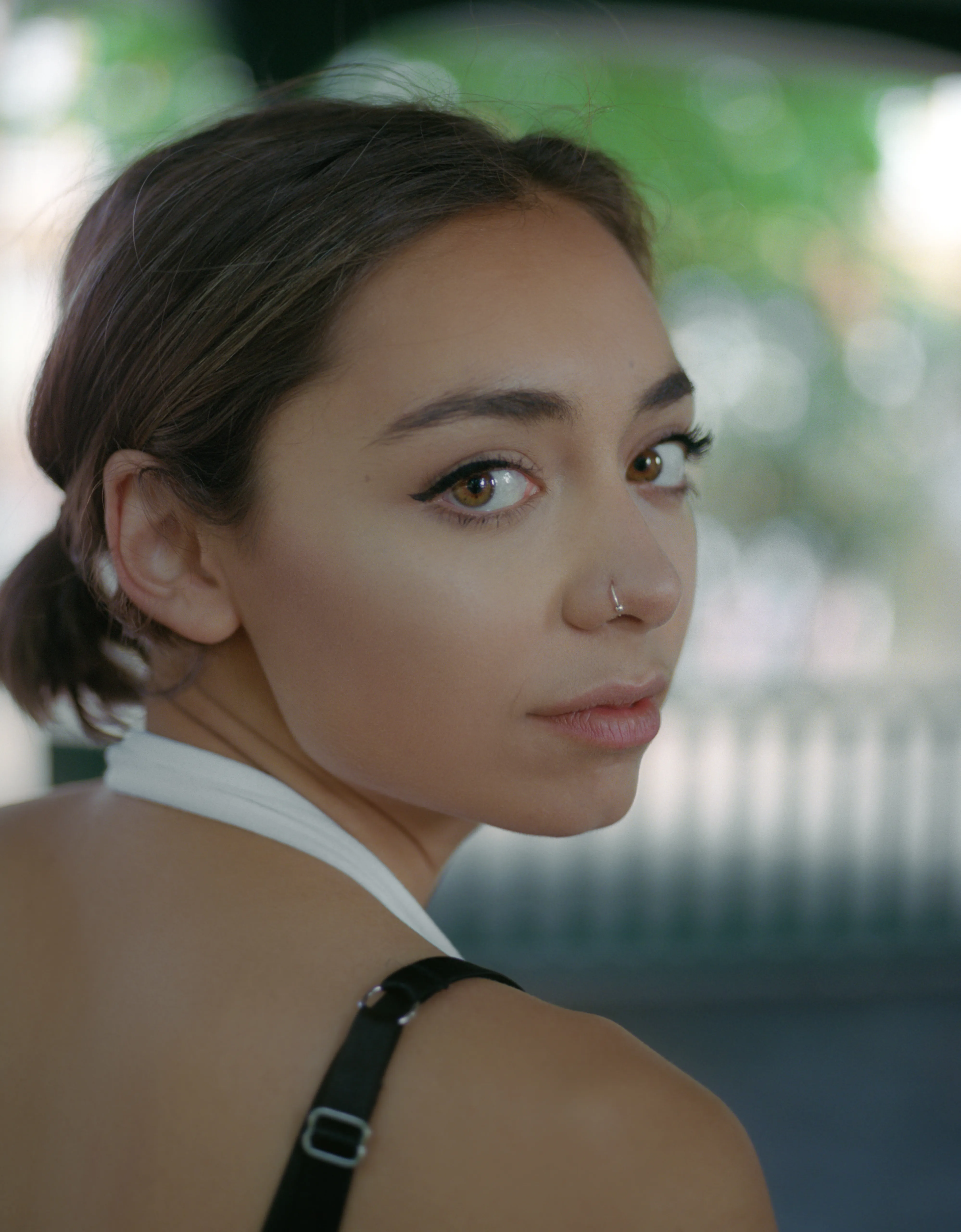 Riya turns to look back over her bare shoulder directly into the camera, her face lit against soft green foliage.