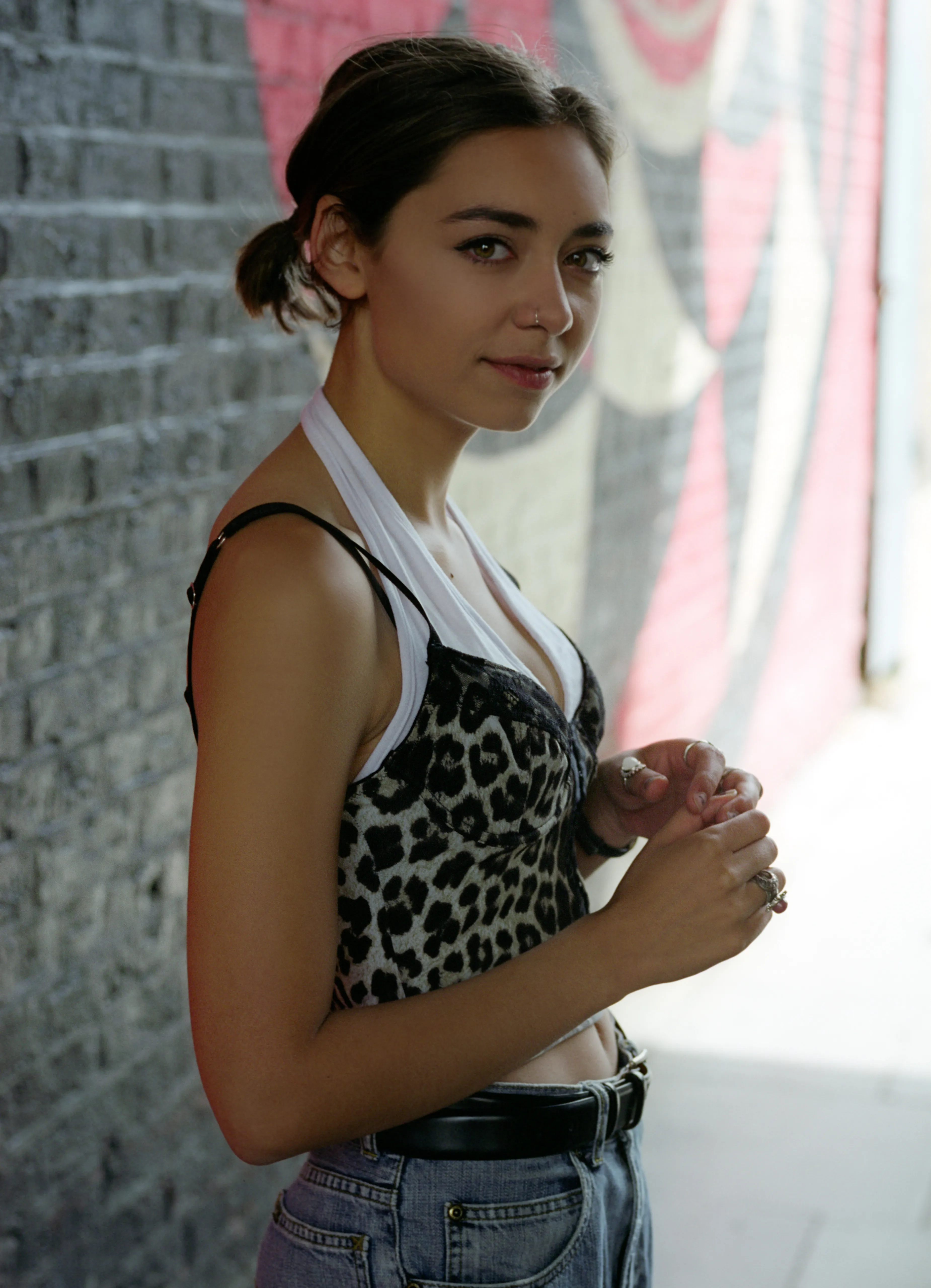 Riya leans against a grey brick wall, half-turned towards the camera with her hands clasped at her waist, a brightly painted mural behind her.