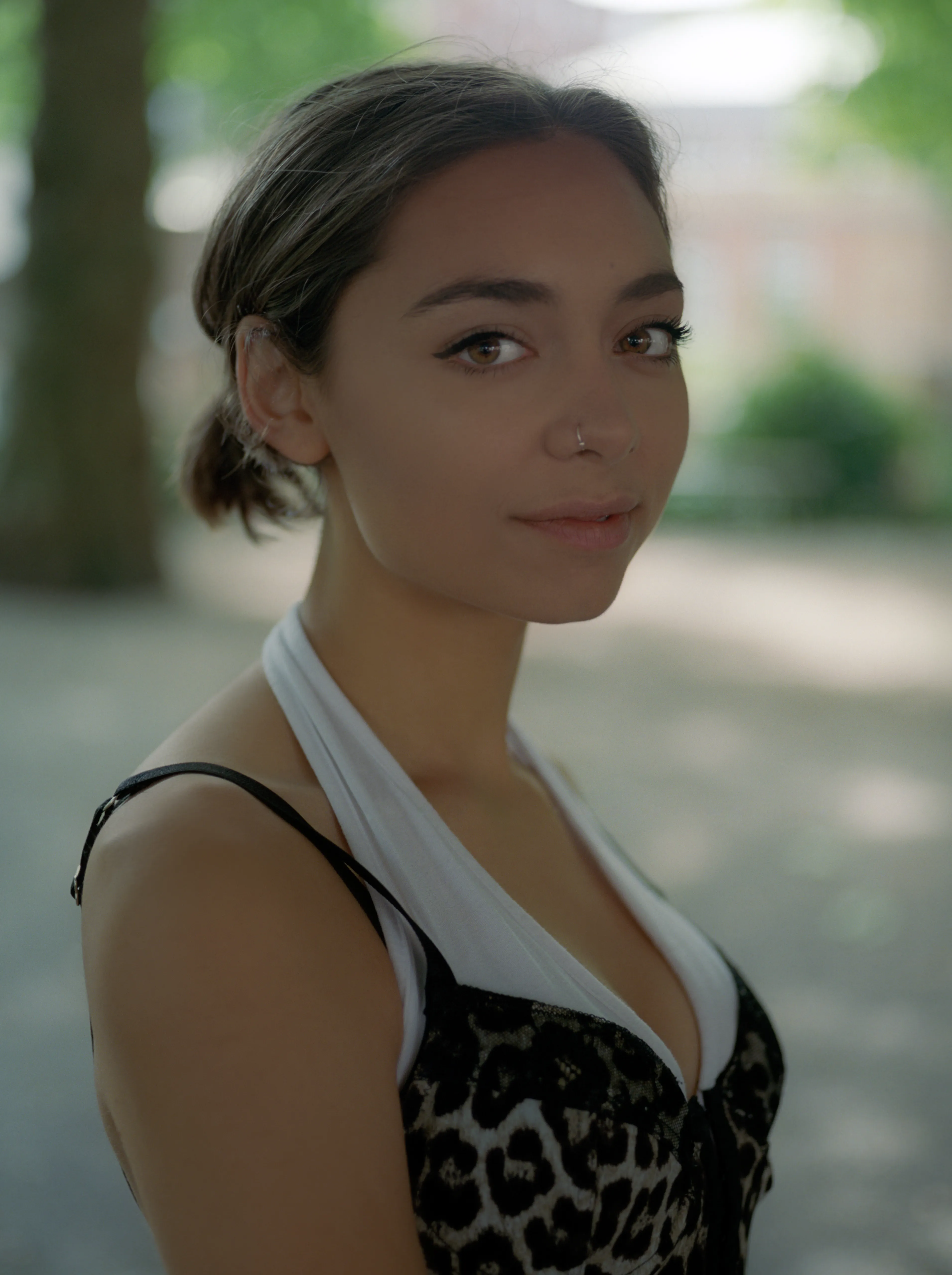 A portrait of Riya in a leafy park, turned three-quarters towards the camera with a calm half-smile, soft green light behind her.